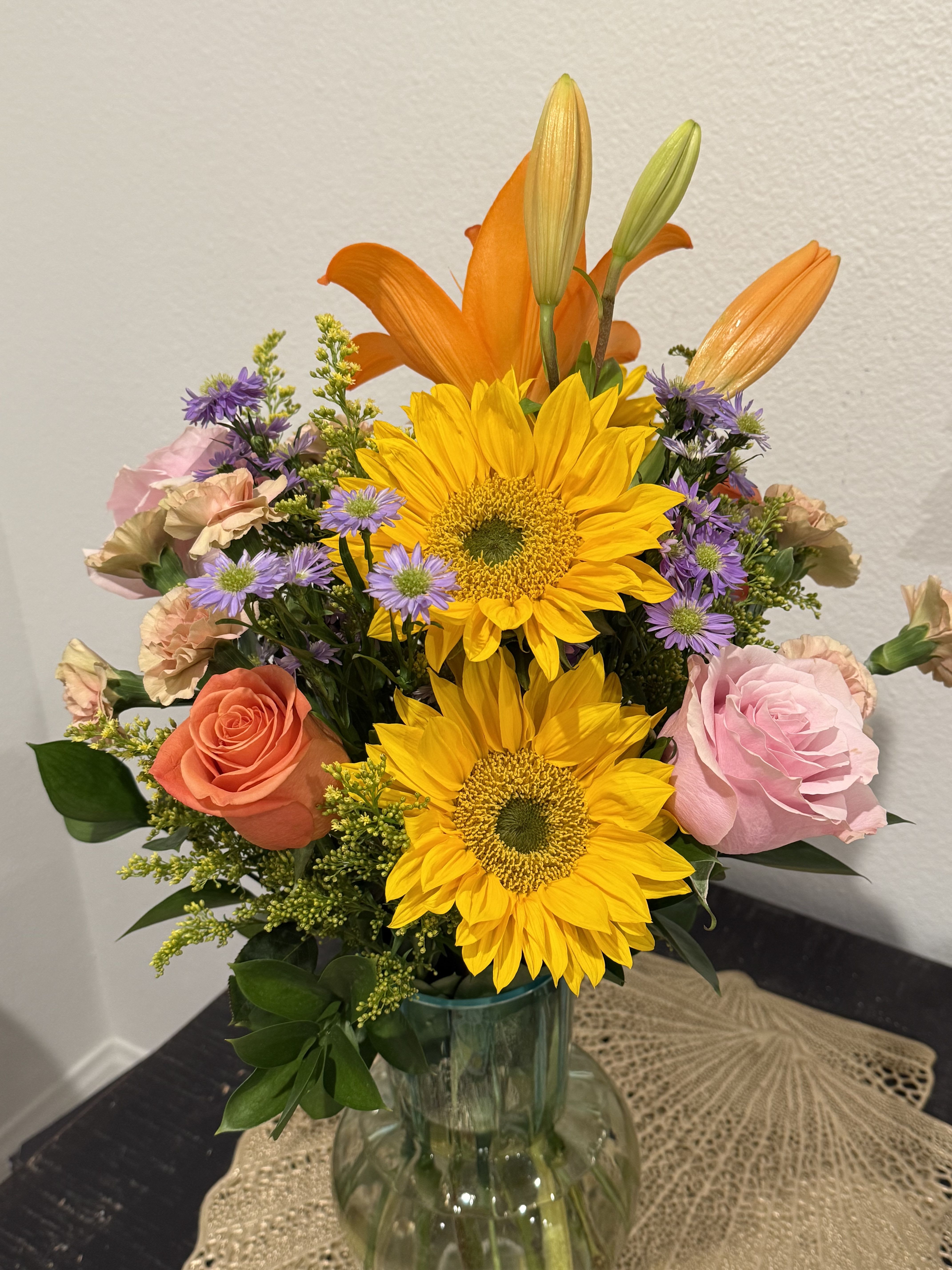 Mixed bouquet with sunflowers, roses, lilies, and purple daisies in a glass vase