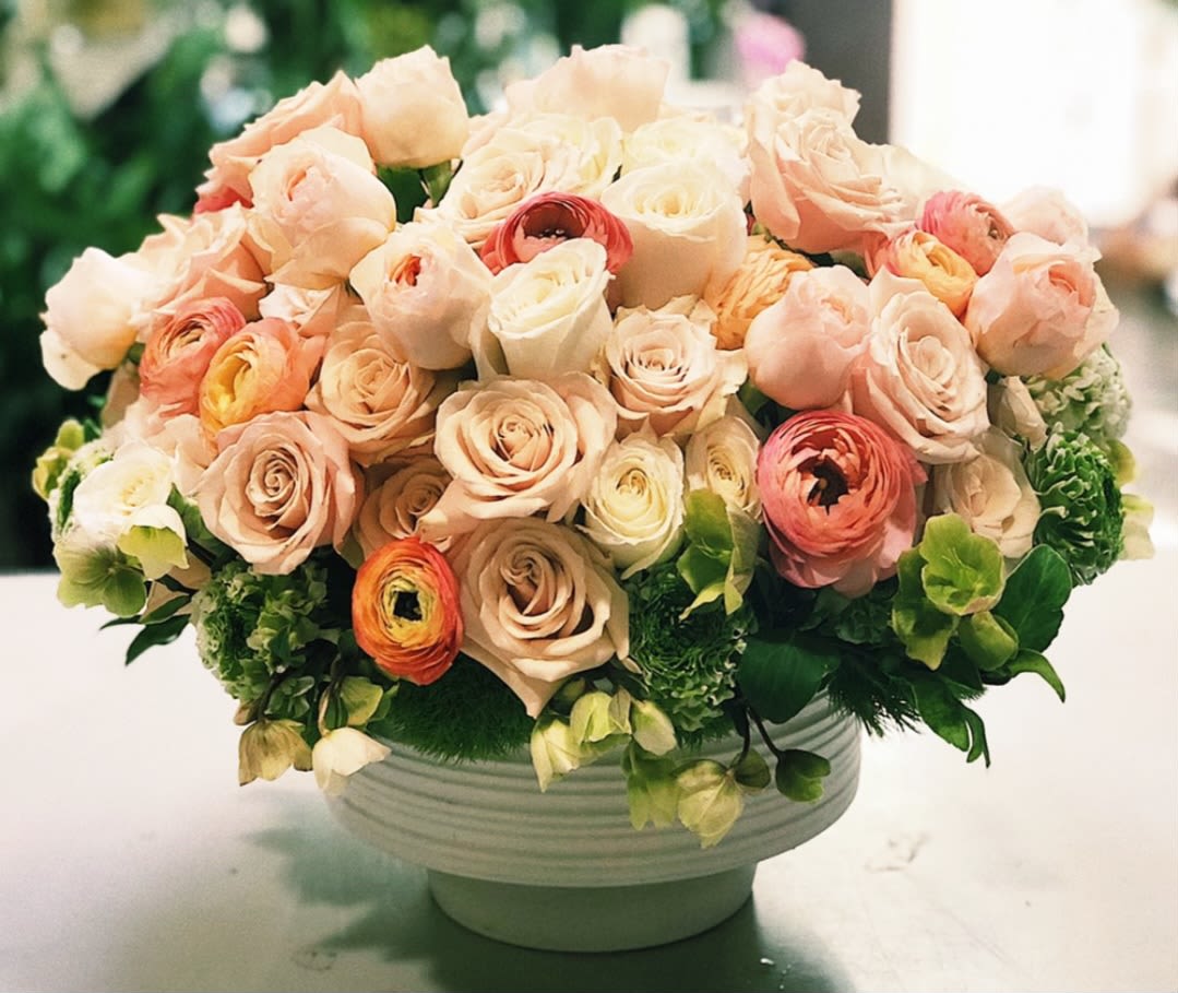Round bouquet of blush and cream roses with green accents in a white bowl vase