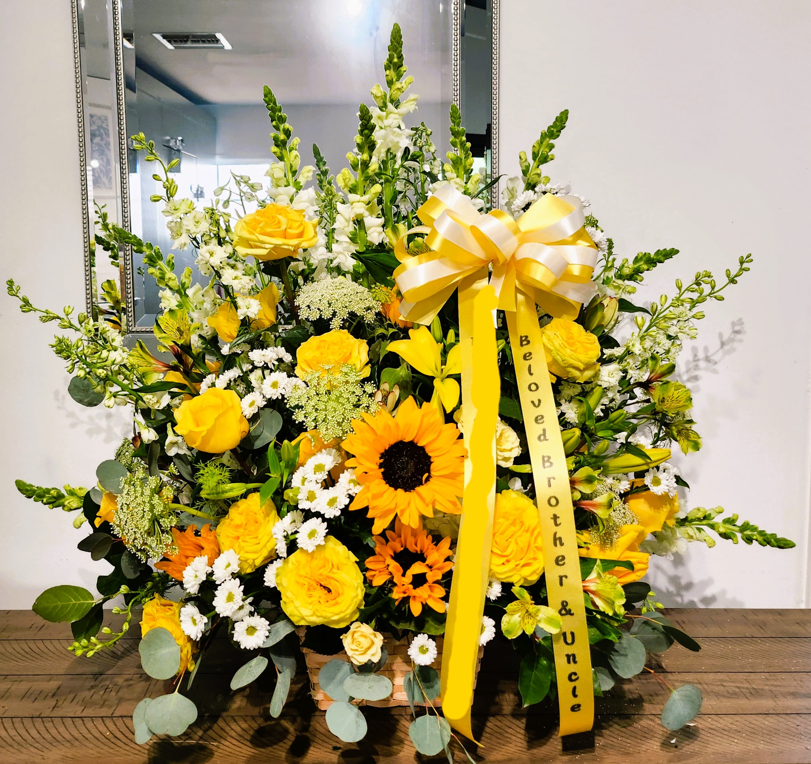 Tall yellow and white sympathy arrangement with roses, sunflowers, and snapdragons in a basket with memorial ribbon.