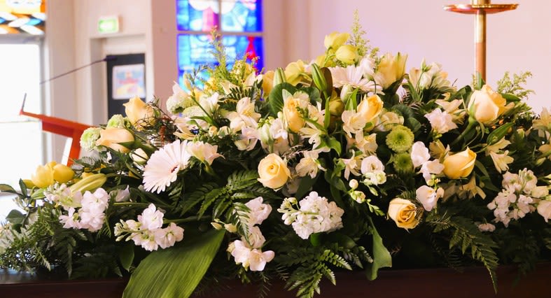 Long arrangement of white and pale yellow roses, lilies, and daisies on a casket in a church setting