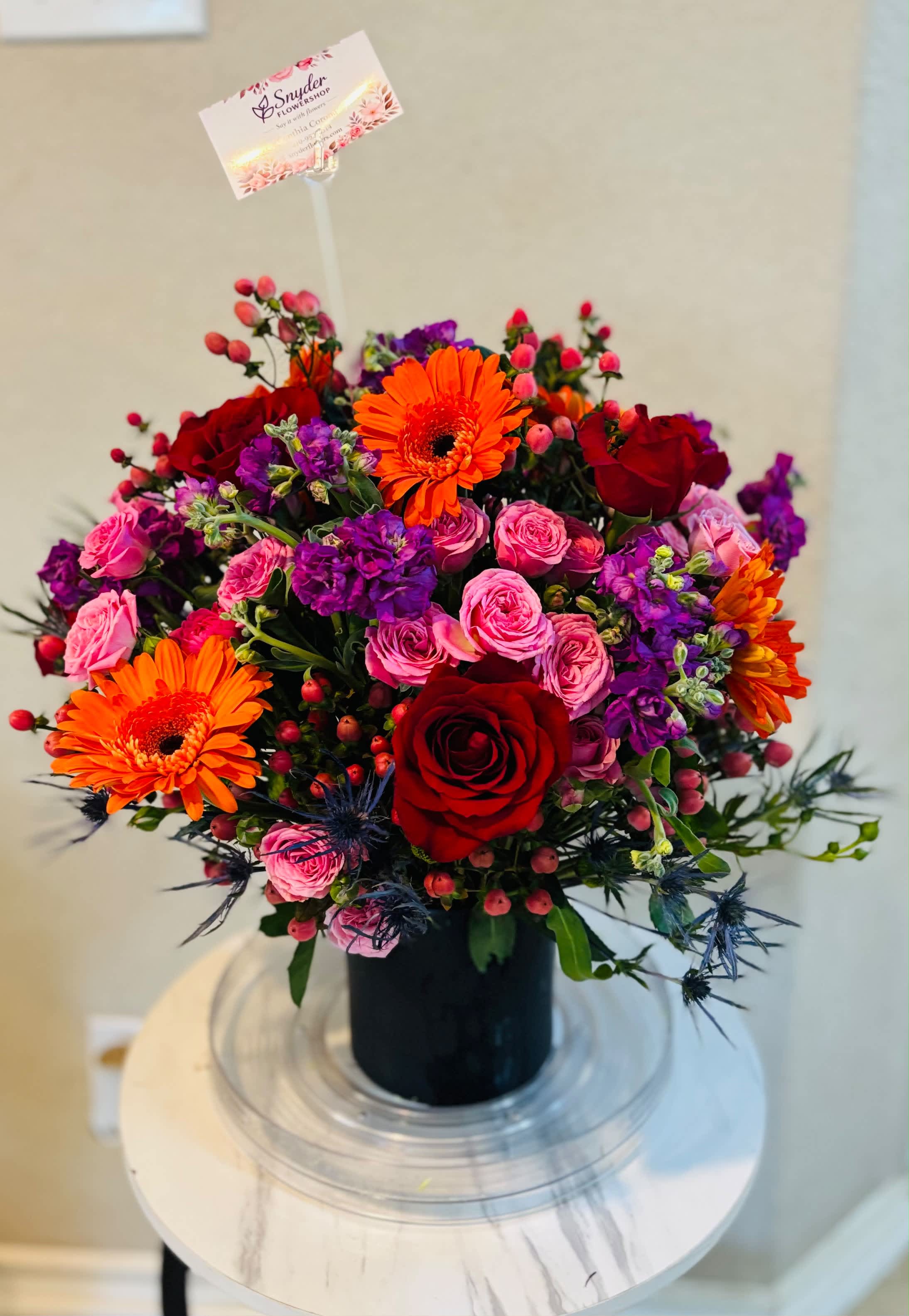 Bright mixed bouquet with red roses, pink spray roses, orange gerberas, and purple blooms in a black vase