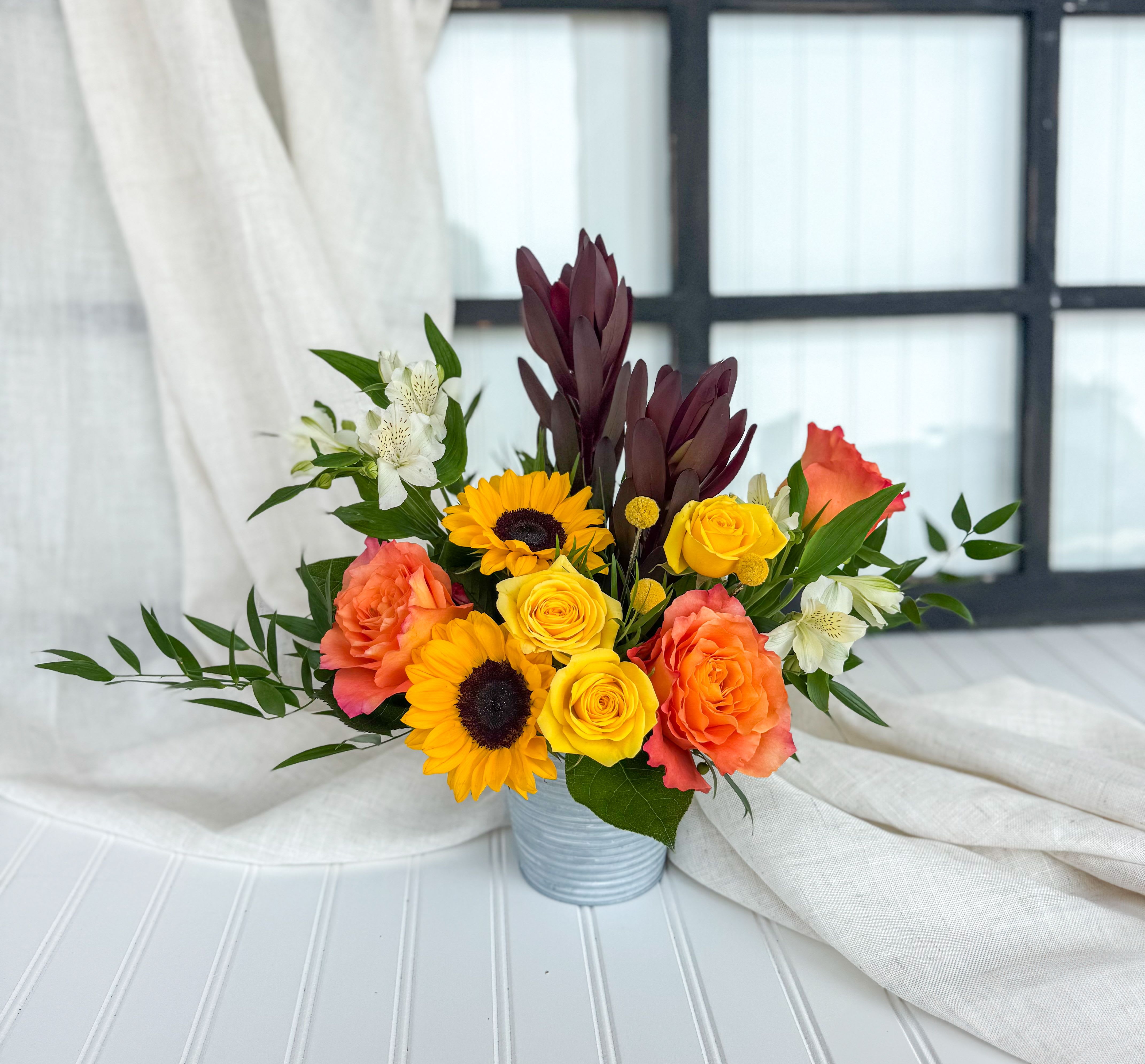 Mixed bouquet of yellow roses, sunflowers, and coral blooms in a metal container
