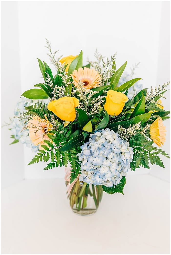 Bouquet of yellow roses, gerbera daisies, and blue hydrangea in a glass vase