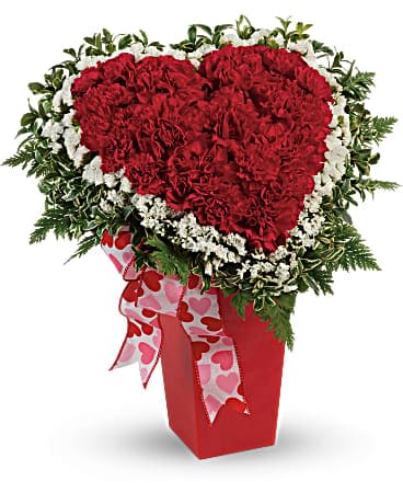 Heart-shaped red carnation arrangement in a red vase with a heart ribbon