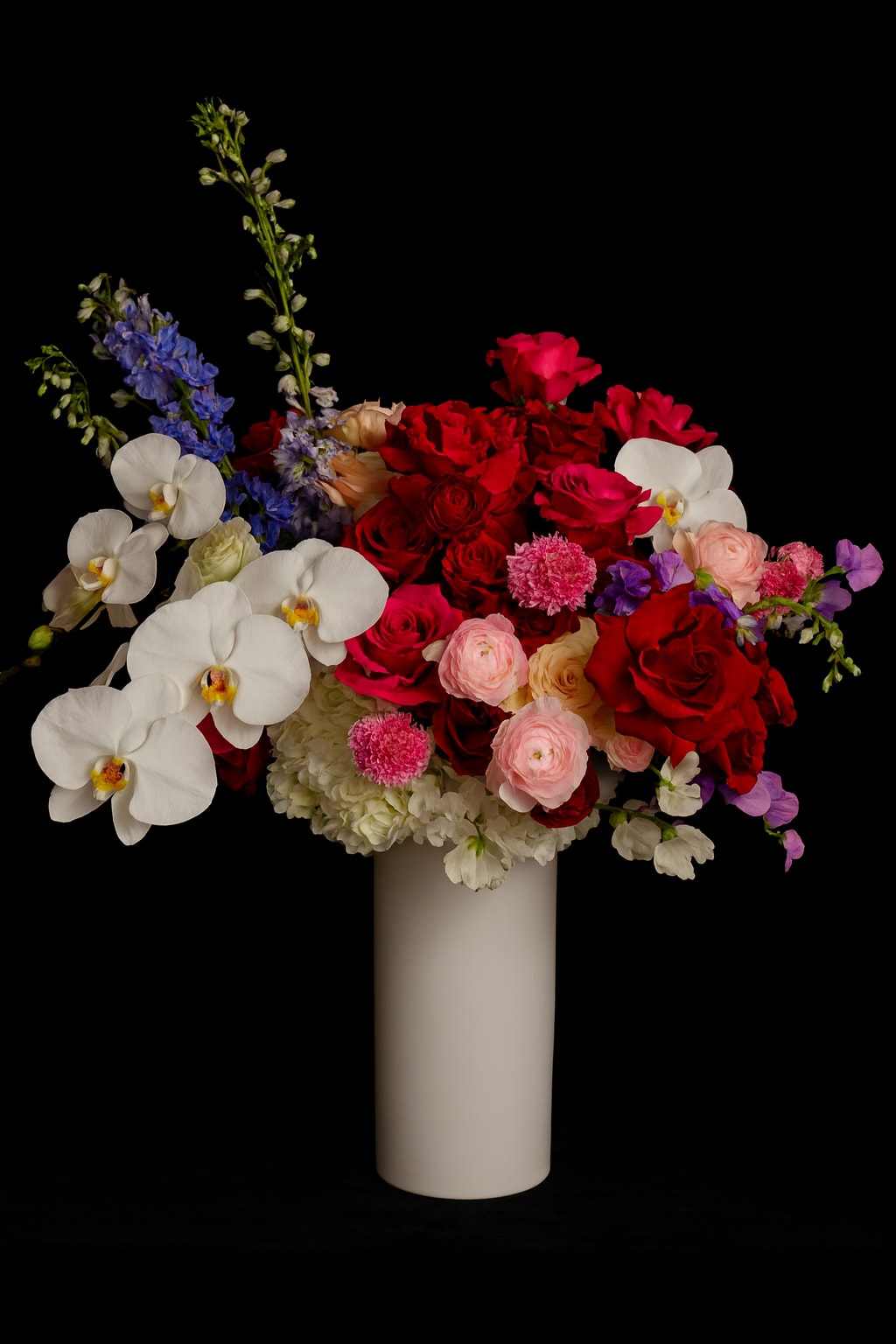 Mixed bouquet of red roses, white orchids, and blue flowers in a white vase