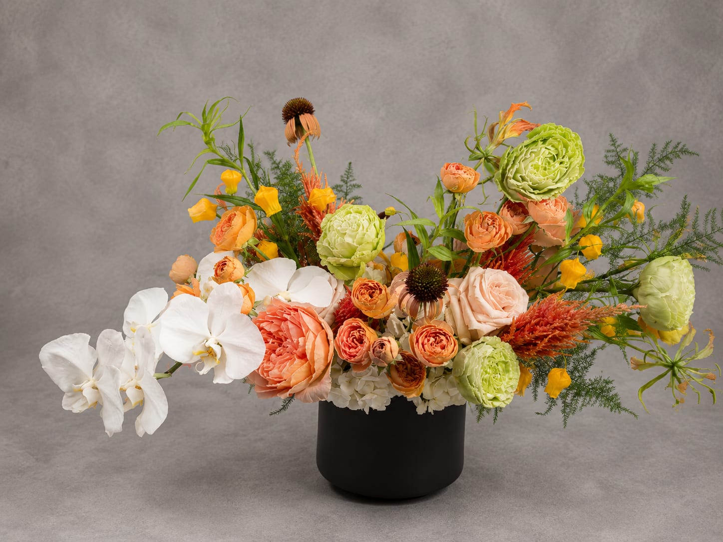 Jessie - Vibrant, graceful, and endlessly inviting, Jessie captures the effortless beauty of a flourishing garden. Layers of apricot garden roses, sculptural orchids, fresh greens, and cheerful seasonal blooms come together in a design that's both refined and full of life. Every stem is thoughtfully placed to create movement, texture, and a sense of natural abundance.