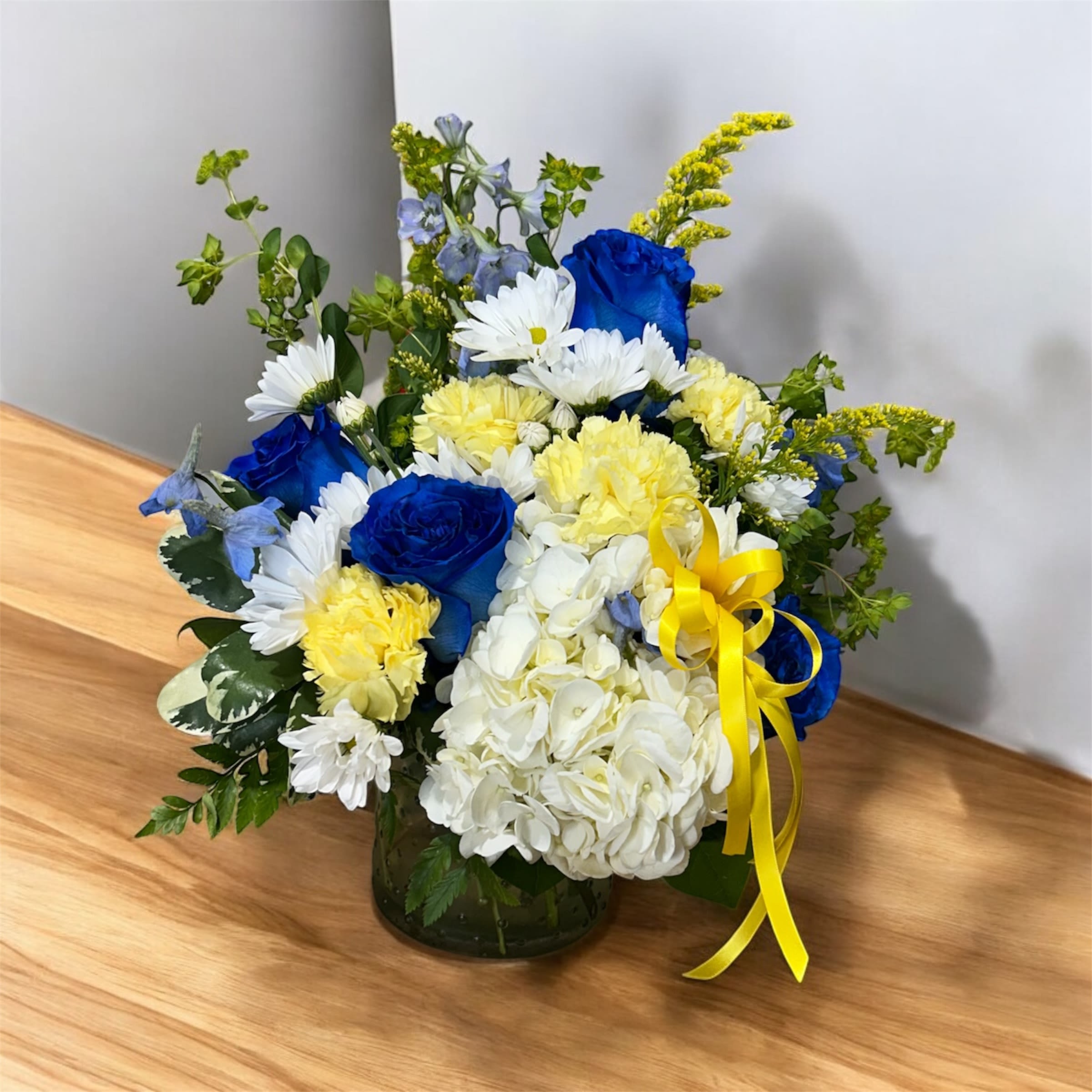 Blue roses, white hydrangea, and yellow flowers in a glass vase with ribbon