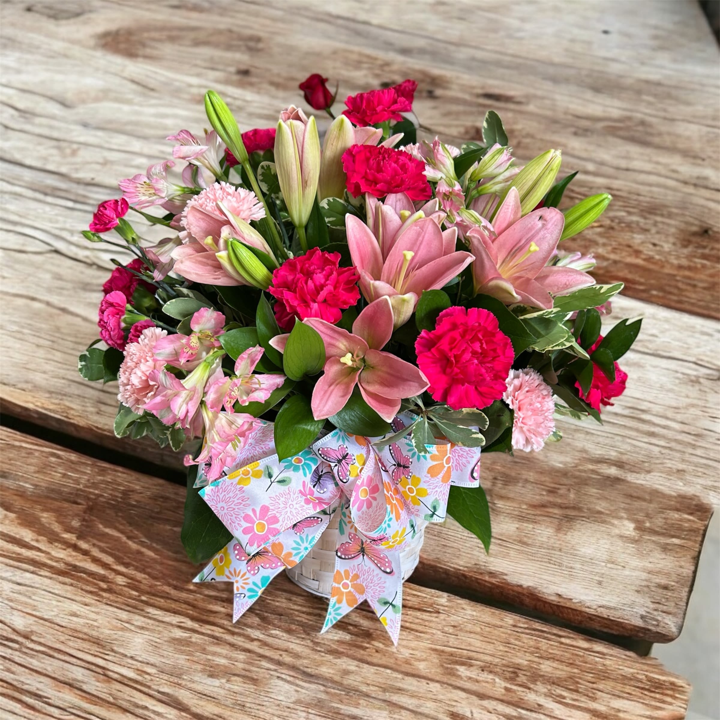 Basket arrangement of pink lilies and carnations with a floral butterfly ribbon.