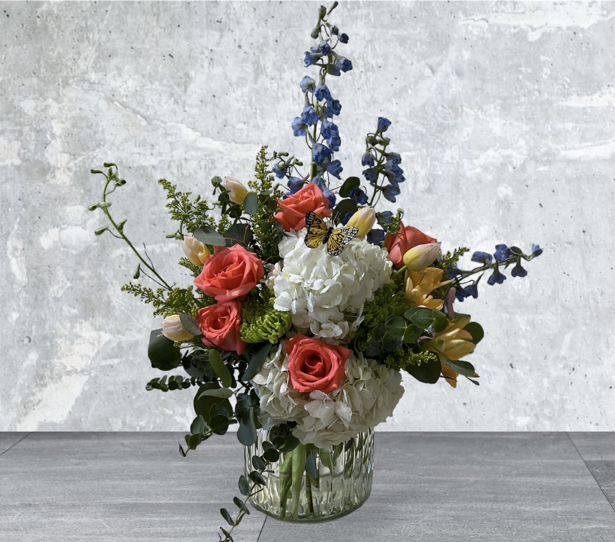 Mixed arrangement of white hydrangeas, coral roses, tulips, and blue delphinium in a clear glass vase
