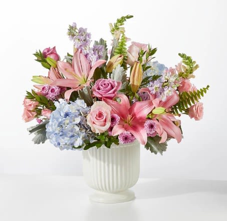 Wonderland Bouquet - A lush, garden-style arrangement of pink lilies and roses with pale blue hydrangea sets the tone for this romantic everyday bouquet-perfect as a birthday bouquet, Mother's Day surprise, anniversary flowers, or a thoughtful "thinking of you" gift. The soft pastel mix of pink, white, blue, and purple reads light and cheerful so the person you're sending it to will feel genuinely celebrated and comforted the moment it arrives.  This full, rounded design is arranged in a white fluted/ribbed ceramic vase that keeps the presentation clean and classic while letting the blooms shine. Expect abundant pink lilies and roses, a generous cluster of pale blue hydrangea near the front, plus lavender-toned stock and dianthus with airy green foliage for a layered, garden-washed texture. The sizeable, centerpiece-like presence makes it suitable for tabletops or entryways and the delicate color palette pairs easily with springtime or everyday décor.  Send it to someone who appreciates soft, romantic florals-the bouquet reads as both refined and relaxed, the kind of arrangement that brightens a room and makes the recipient feel truly seen. Vase-arranged and ready to display; keep flowers fresh by topping up water daily and trimming stems every few days.