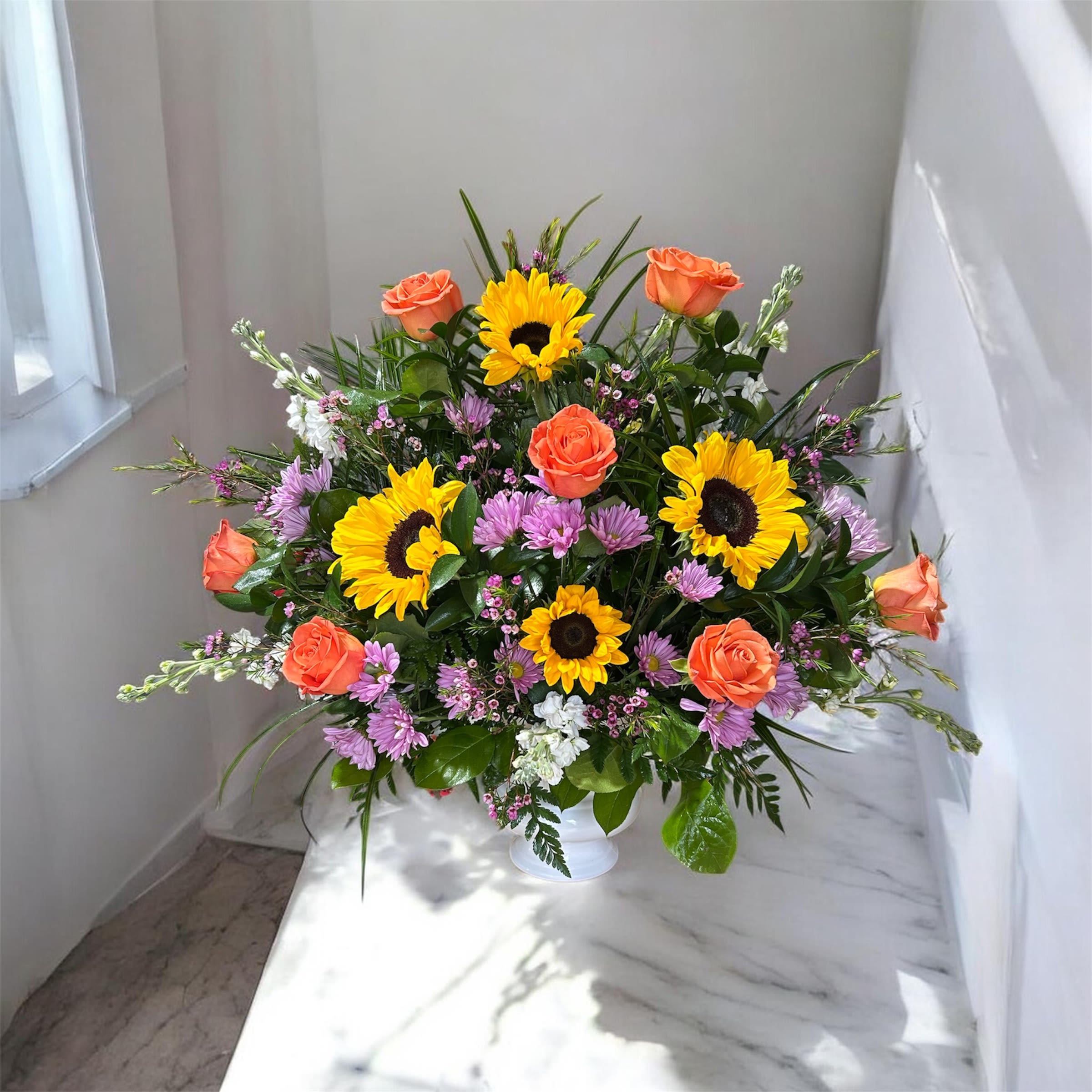 Cheerful Goodbye  - Big, golden sunflowers take center stage in this cheerful birthday bouquet, paired with warm orange roses and lively purple daisy-like clusters for a bright, celebratory look that makes the recipient feel instantly uplifted. The mix of sunny yellow, tangerine and cool lavender tones creates a joyful contrast perfect for birthdays, congratulations, or "thinking of you" moments when you want someone to feel truly celebrated.  Arranged in a clean white ceramic vase, the design reads full and rounded with a relaxed garden-style texture-soft rose petals, bold sunflower faces, and peppered small blooms that add playful movement. Colors: yellow, orange, purple and white. Flowers include sunflowers, roses, daisies and complementary filler blooms; vase included for easy display. The scale and fullness give an immediate table-ready presence without fuss.  This is the kind of bouquet that brightens a kitchen island, office desk, or bedside table and sends a sunny, optimistic message to the person you're thinking of. Order with standard delivery to ensure peak freshness; simple care-fresh water and a cool spot-keeps blooms cheerful for days.