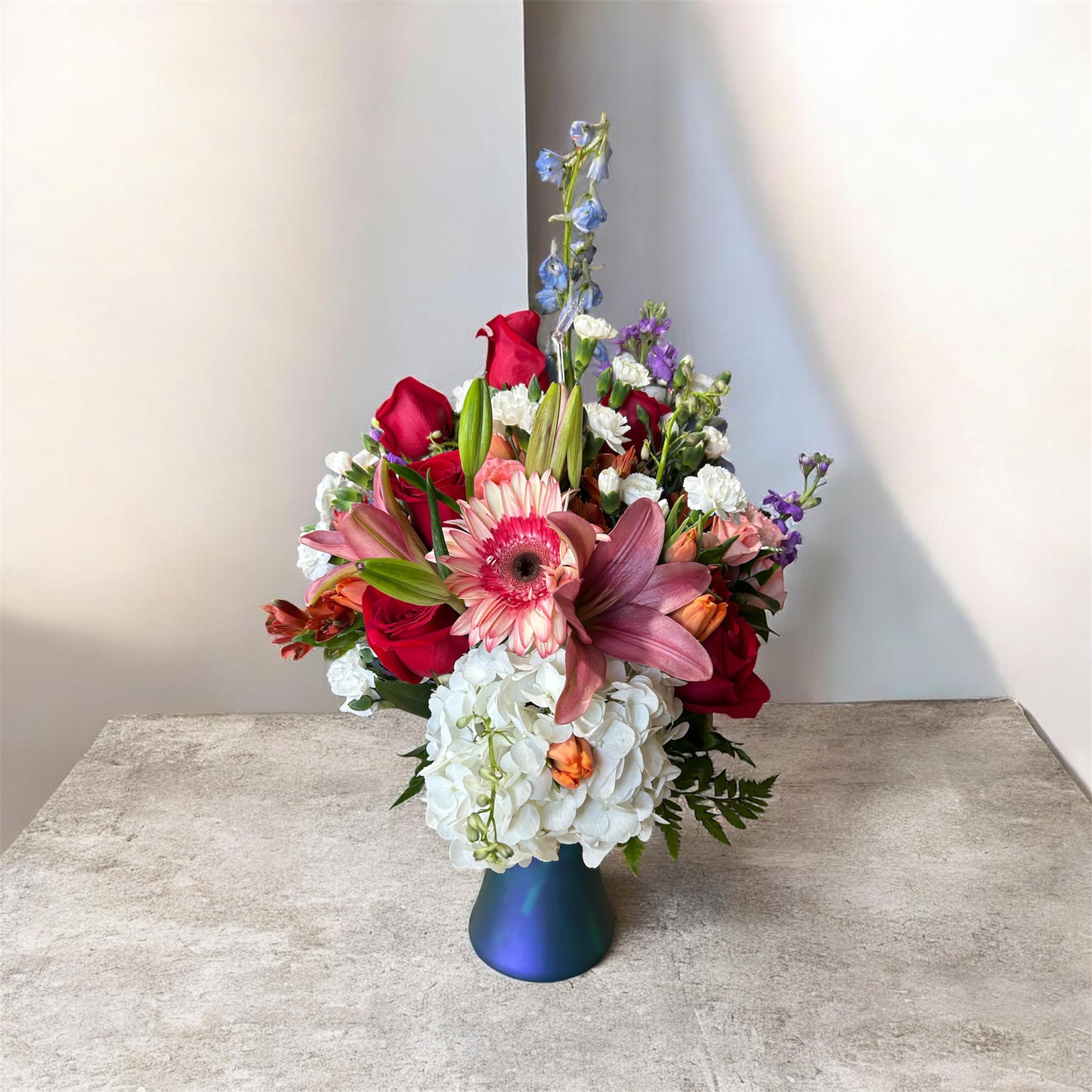 Cheerful Day - A vivid, romantic mix that catches the eye-Cheerful Day is a tall, lush flower arrangement perfect as a birthday bouquet, anniversary gift, or romantic surprise. The composition reads bright and celebratory: the vertical blue spike rises above a dense cluster so the person you're thinking of will feel truly noticed the moment it's set down.  This arrangement features red roses, pink and purple lilies, a pink gerbera daisy, white hydrangeas and delicate white and purple filler blooms layered tightly for a full, textured look. Stock and alstroemeria add fragrance and small-flower detail while the soft white hydrangeas balance the vivid pinks, reds and purples. Delivered in a blue vase, the piece has a compact footprint with graceful height from the central spike-ideal for a mantel, dining table centerpiece, or anywhere you want color and presence.  Send this when you want someone to feel celebrated and cherished-whether marking a milestone, saying "I love you," or brightening their day. Standard care: trim stems and refresh water every few days to keep the blooms looking their best.