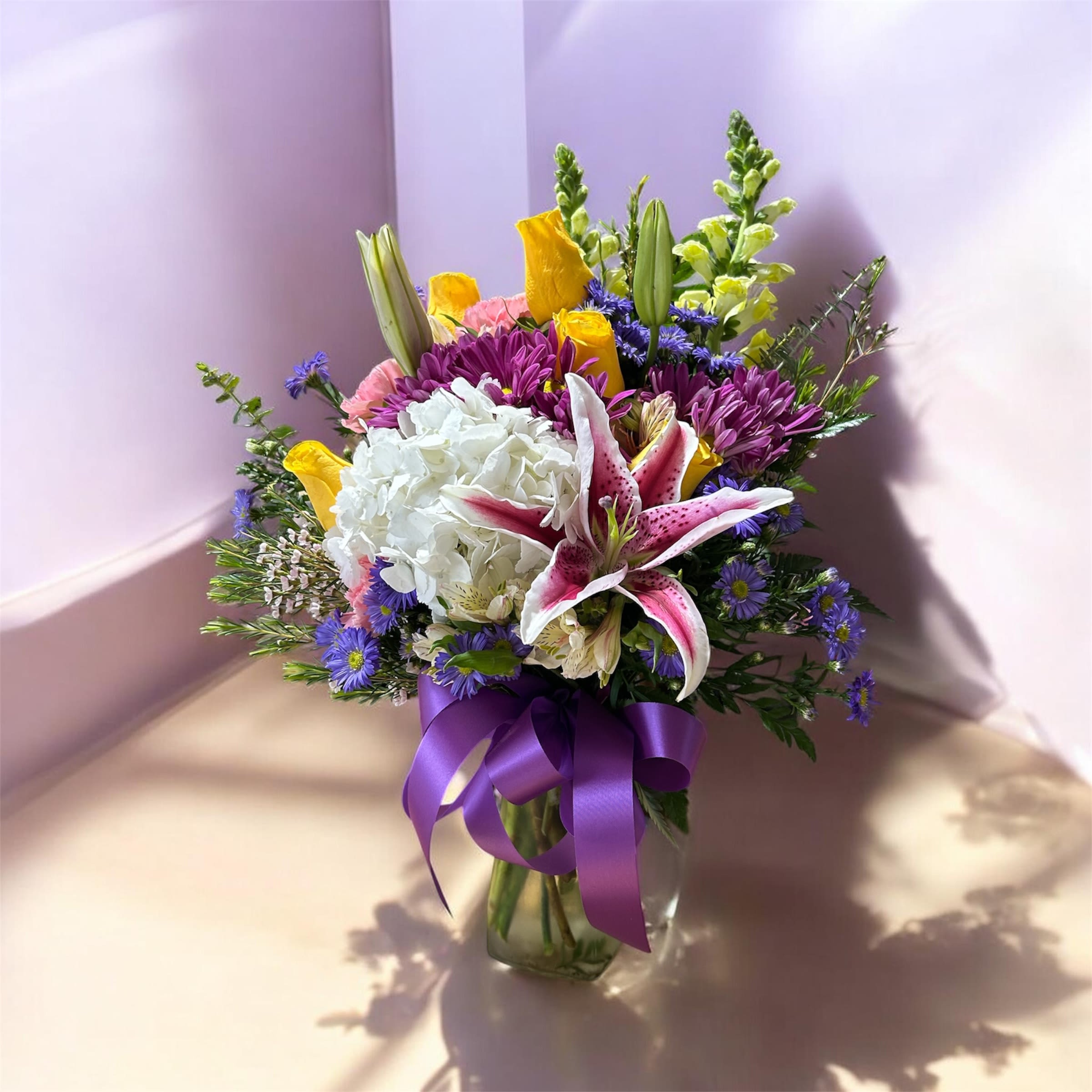 Enchanted Garden Bouquet - A lush cluster of white hydrangea anchors this Enchanted Garden flower arrangement, surrounded by pink-and-white lilies, purple daisy-like blooms, sunny yellow accents, and soft chrysanthemums for a garden-fresh feel - perfect as a birthday bouquet or a cheerful anniversary surprise. The rounded, layered composition and pops of purple make the recipient feel seen and celebrated, whether you're marking a milestone or simply saying "I'm thinking of you."  Arranged in a clear glass vase with a wide purple ribbon tied around the neck, the bouquet reads bright, romantic, and effortlessly elegant. Taller buds and spike elements rise above the main body to create depth and movement; textures range from the hydrangea's cloudlike softness to the lilies' sculpted petals and the daisies' playful centers. The size and fullness make it an impressive centerpiece for a dining table or a welcoming gift to leave at a doorstep.  This is the kind of bouquet that fits many moments - from congratulations to get-well wishes - and the clear vase keeps the look clean and modern. Change the water every two days, trim stems slightly when refreshing, and remove any faded blooms to extend enjoyment; available for same-day local delivery when ordered before cut-off.