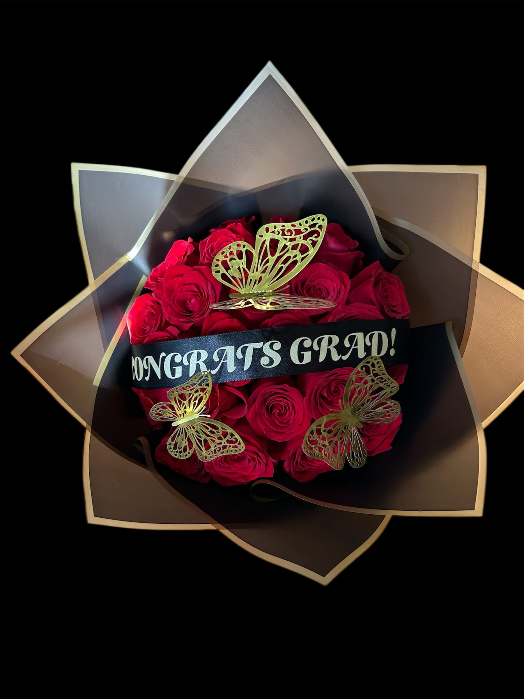 You Did It Bouquet 24 Stems - A bold cluster of vivid red roses makes a confident graduation bouquet-perfect as a graduation flower arrangement to celebrate commencement or academic milestones. The compact, decorative presentation reads youthful and celebratory: the red-and-black contrast catches the eye and the wide black ribbon that says "CONGRATS GRAD!" ensures the recipient instantly knows this is their moment.  Wrapped in layered black and translucent geometric paper, the roses are accented with three metallic butterfly ornaments that add sparkle and motion against the velvety petals. The arrangement is finished with a black ribbon across the center for a polished, gift-ready look. Ideal for gifting to a graduate who loves dramatic color and modern style, this bouquet arrives ready to display and makes someone feel distinctly celebrated.
