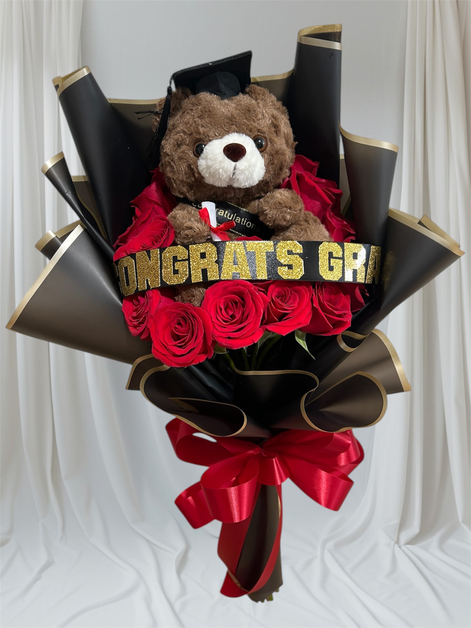 Cheerful Grad - A cheerful graduation bouquet built around a plush brown teddy bear in a mortarboard makes a playful, celebratory statement for the new graduate. This graduation bouquet pairs classic red roses with bold black decorative wrapping and a glittery congratulatory band so the recipient instantly knows the moment is being honored-the kind of gift that reads "congratulations" before they even open it.  Rich red roses surround the cap-wearing teddy, accented by white and pink highlights visible among the blooms and finished with a red satin bow at the base. The arrangement is presented in dramatic black paper with gold trim and tied with ribbon, giving a polished, photo-ready look that holds up for gifting or campus photos. Glitter text on the band adds a festive sparkle without overpowering the roses.  Perfect for commencement day, a celebratory send-off, or to mark academic milestones, this bouquet is geared to make the graduate feel seen and celebrated. Order with standard sizing as shown; stems are arranged for easy display and simple care-keep flowers in fresh water, out of direct sun, and enjoy the teddy keepsake long after the blooms have faded.