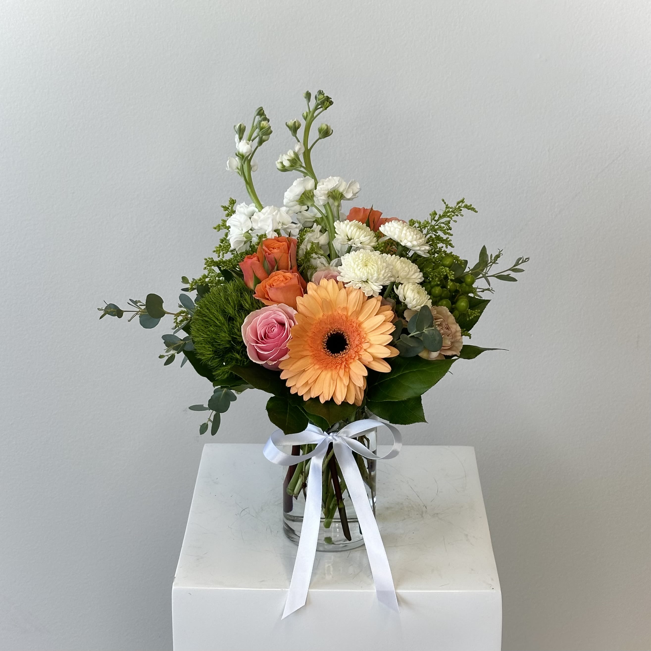 Sweet Tea - Sweet Tea is a heartwarming floral embrace that evokes lazy summer afternoons and the gentle comfort of a tall glass of iced tea kissed by sunshine. This radiant arrangement blends warm peach and vibrant orange tones with soft white accents, creating a welcoming, joyful display full of texture, fragrance, and effortless charm. A bold orange gerbera daisy takes center stage, its wide, sunny petals beaming with optimism and energy. Elegant peach roses nestle alongside, their velvety layers revealing classic beauty. Fluffy peach carnations add plush fullness and softness, their soft ruffled petals enhancing the arrangement’s inviting warmth. Playful dianthus balls dot the design with rounded bursts of complementary color, while delicate clusters of orange spray roses weave throughout. Crisp white mums provide a clean contrast and keep the palette fresh and balanced. Graceful stems of white stock rise elegantly and their signature sweet-spicy fragrance lingers beautifully. Wispy golden solidago threads throughout like strands of captured sunlight, its feathery sprays tying the warm hues together with a natural, wildflower-inspired finish. Perfect for birthdays, thank-you gestures, get-well wishes, or simply brightening any space, Sweet Tea delivers uplifting energy and cozy contentment. Its harmonious blend of sunset peaches, fiery oranges, and refreshing whites feels both nostalgic and timeless—ideal for a kitchen island, bedside table, or cozy reading nook. 
