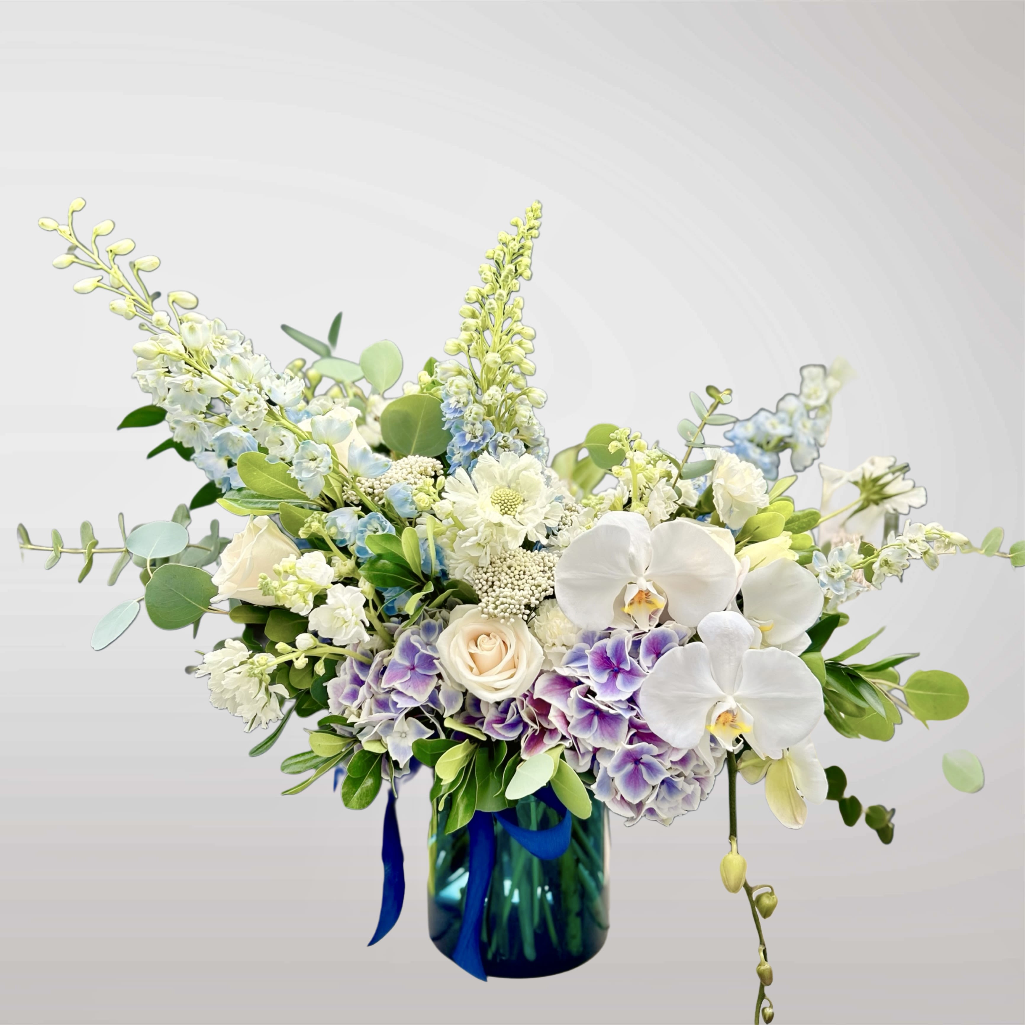 Partly Cloudy - Like a sky after rain, Partly Cloudy drifts between softness and light. Cool blues, creamy whites, and violet tones gather in a lush cloud of blooms, accented with airy textures and elegant orchids. A moment of calm and wonder, captured in flowers.
