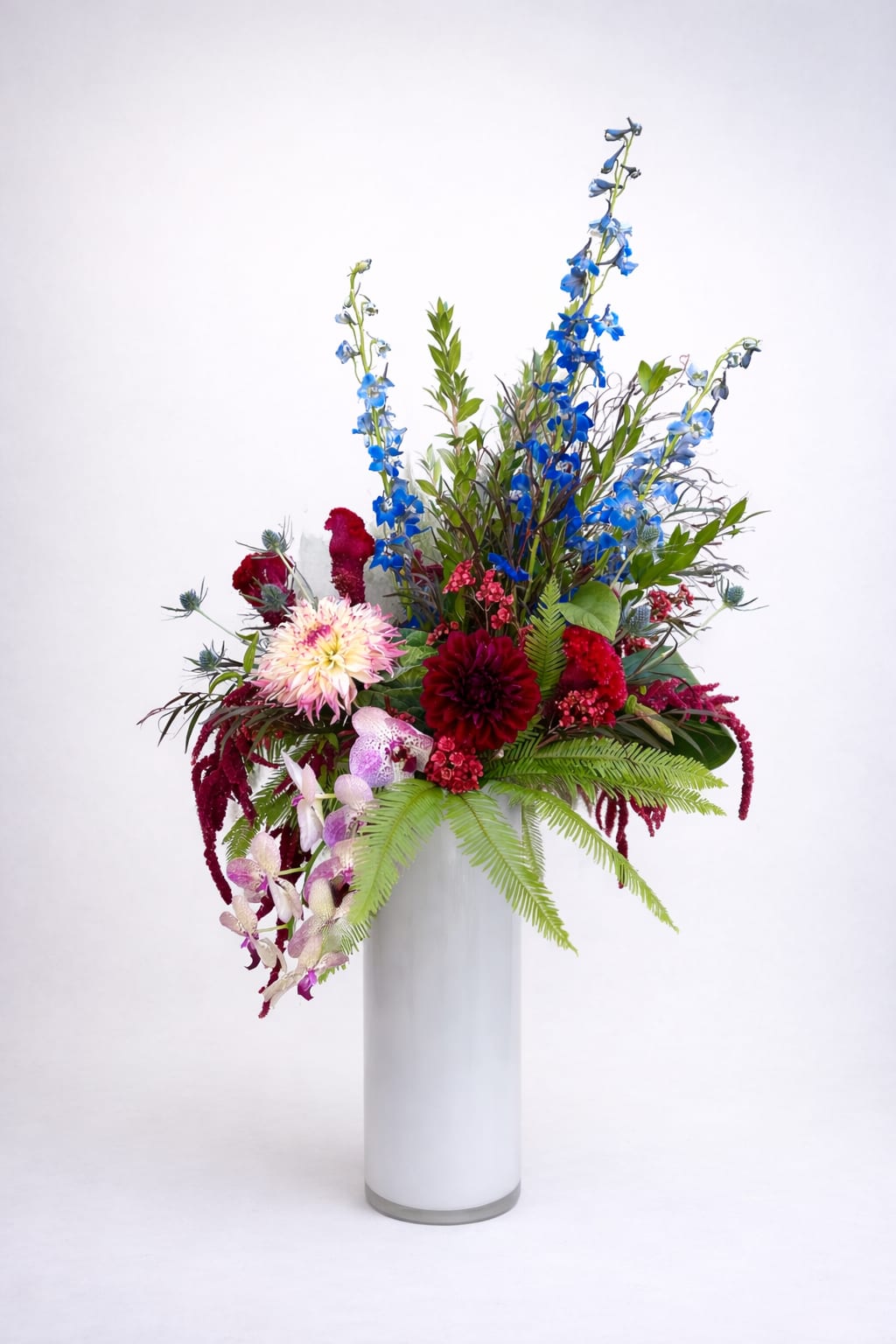 Siren Garden - A mesmerizing symphony of color and motion, Siren Garden captures the magnetic allure of deep waters and untamed blooms. Vivid cobalt delphinium rises like song over a sea of velvety dahlias, orchids, and amaranthus—each petal a note in a haunting melody. Soft ferns unfurl beneath, grounding the piece in lush greenery that whispers of hidden groves and moonlit tides.  Both mysterious and luxurious, this arrangement feels alive with contrast—cool blues against crimson fire, elegance entwined with wild abandon. Siren Garden is a floral aria, calling to the senses with irresistible beauty and a touch of danger, like a secret too lovely to resist.