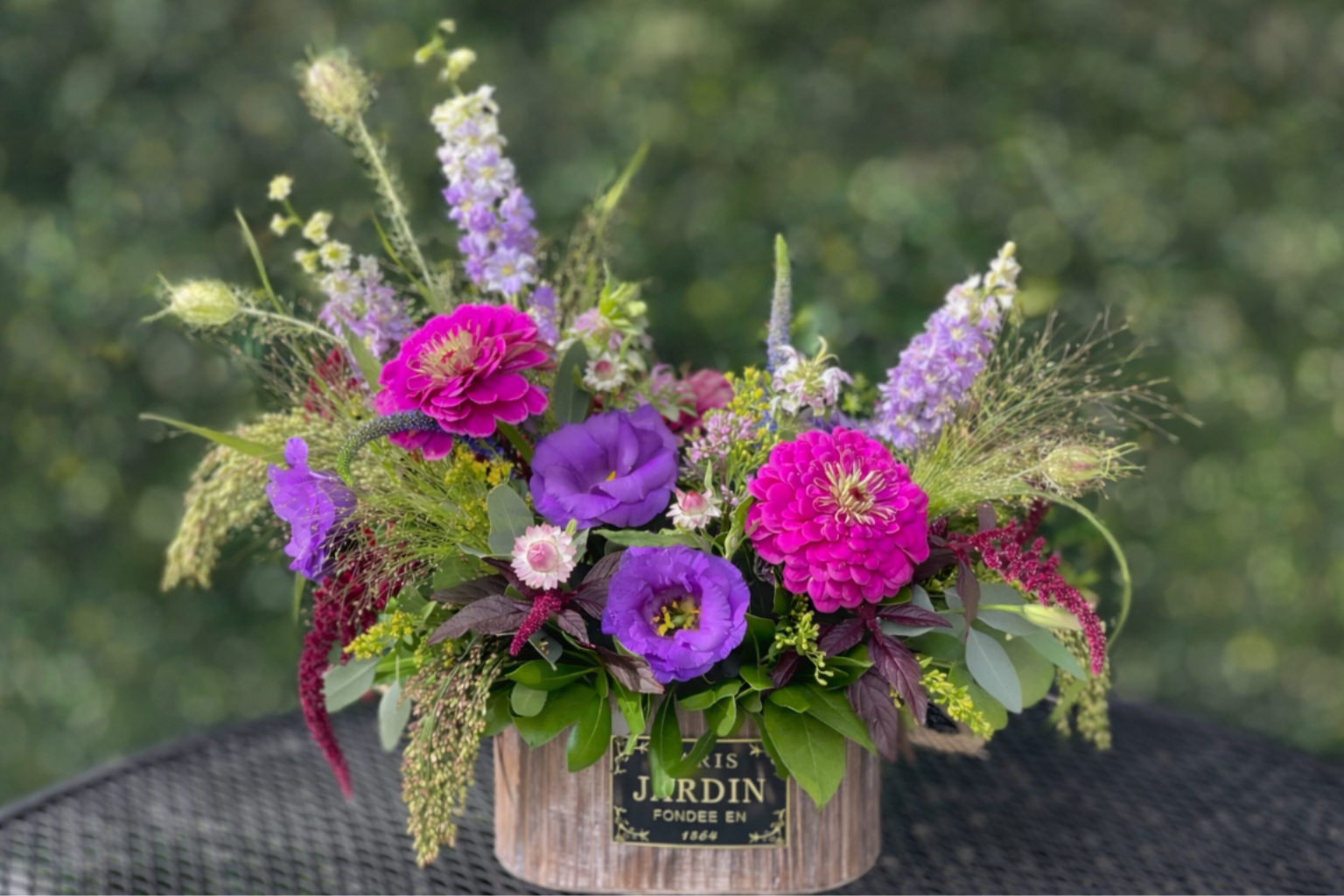 Parisian Prairie - Transport yourself to the French countryside with our Parisian Prairie floral arrangement. This textural assortment evokes an explosion of wildflowers with a mix of fresh blooms, including purple lisianthus, fuscia zinnias, larkspur, burgundy amaranthus, and dynamic grasses, presented in a rustic wooden box.