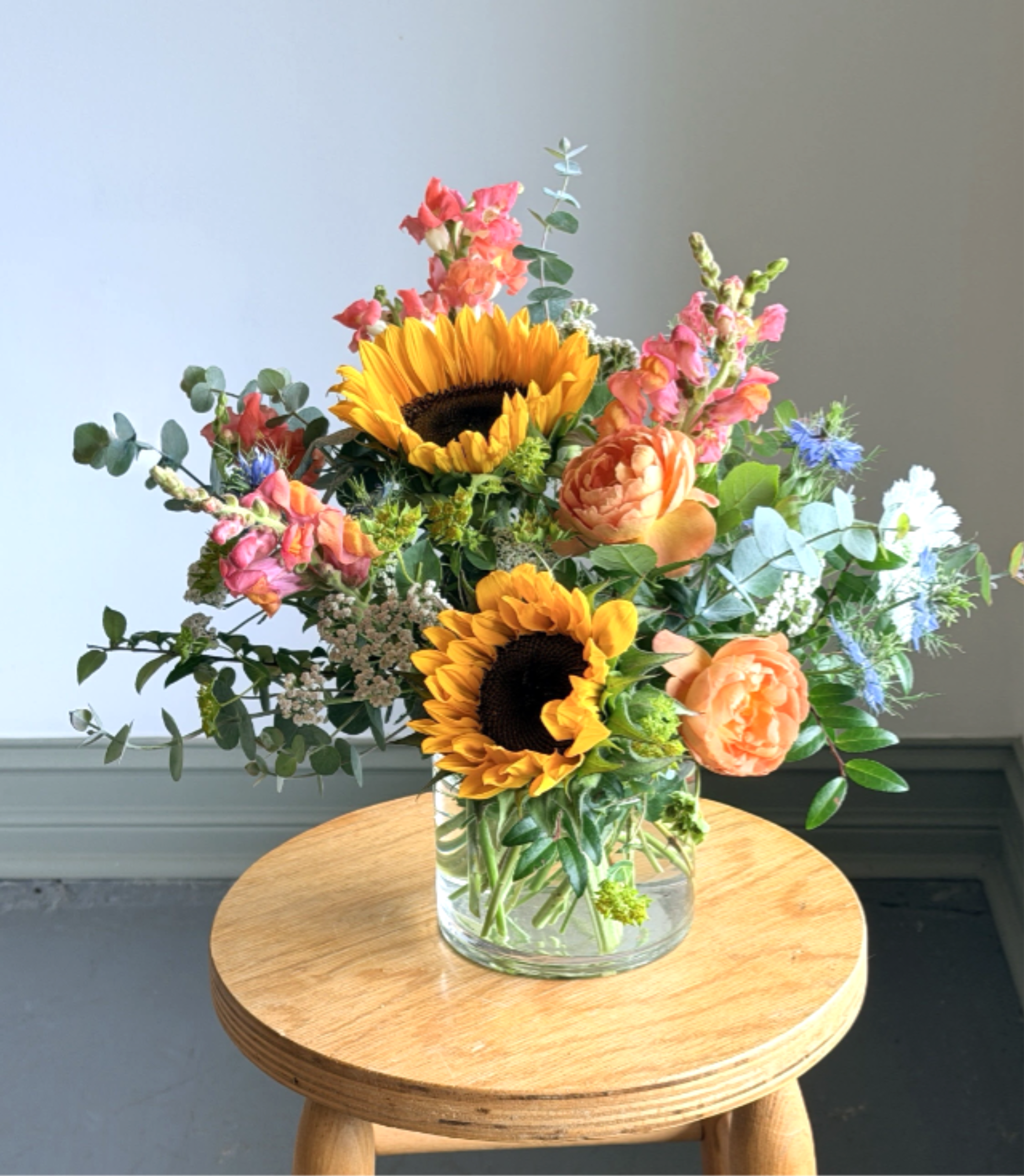 Summer Petite - Brighten their day with bright summer blooms. This petite bouquet is arranged in a glass or ceramic cylinder vessel featuring flowers like sunflowers, lisianthus or garden roses. Always a favorite this arrangement makes a great gift for any occassion.  Want to send something a bit more soft? Let us know you'd like to have this arranged in pastels in the notes section of the order form.  All of our flowers are seasonal, locally sourced and American grown. This allows you to receive the freshest flowers possible for a longer lasting bouquet or arrangement. Each bouquet or arrangement produced is unique to the season and may contain different flowers than what are listed here.