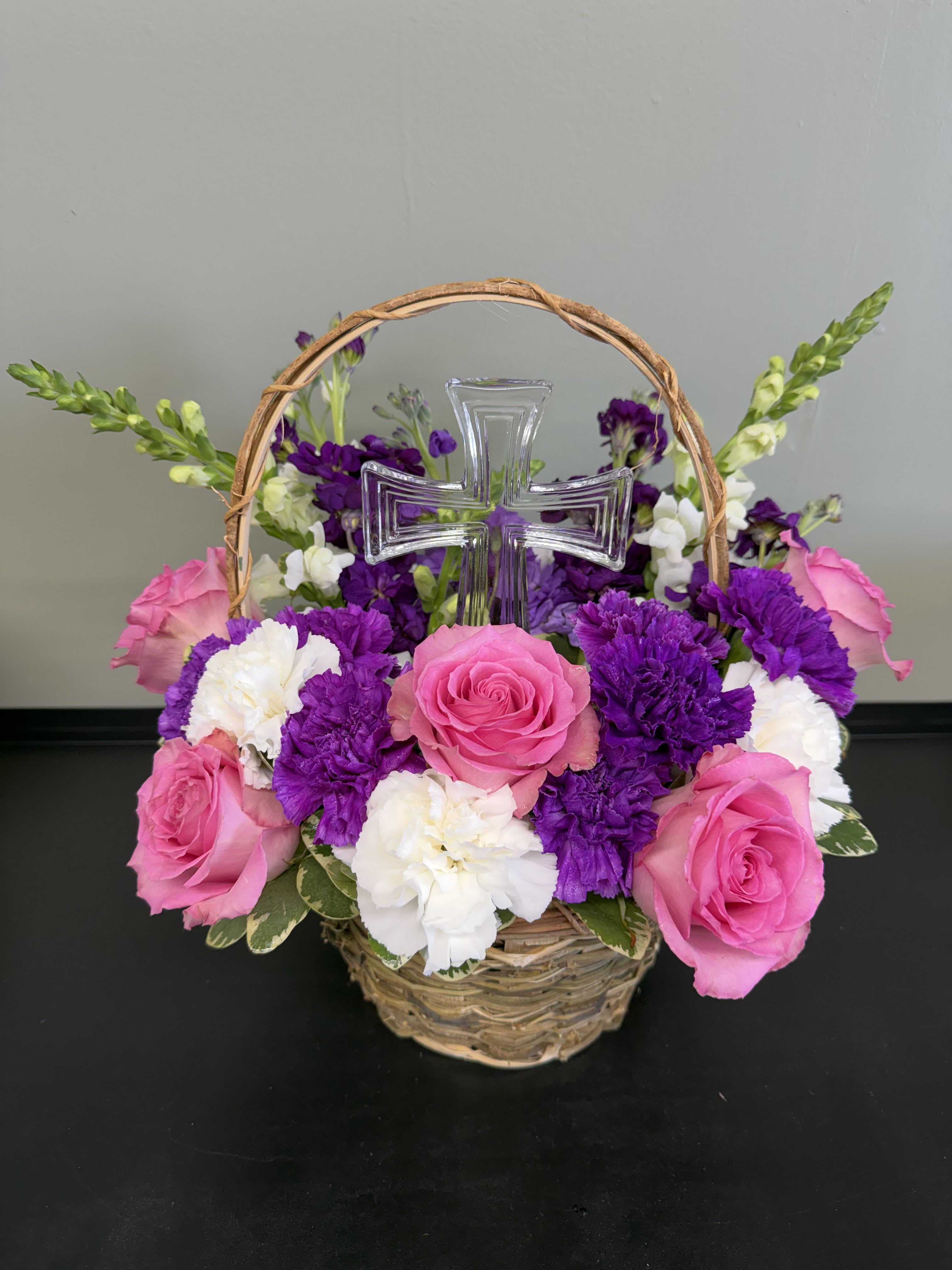 Basket arrangement of pink roses, white carnations, and purple flowers with a clear cross