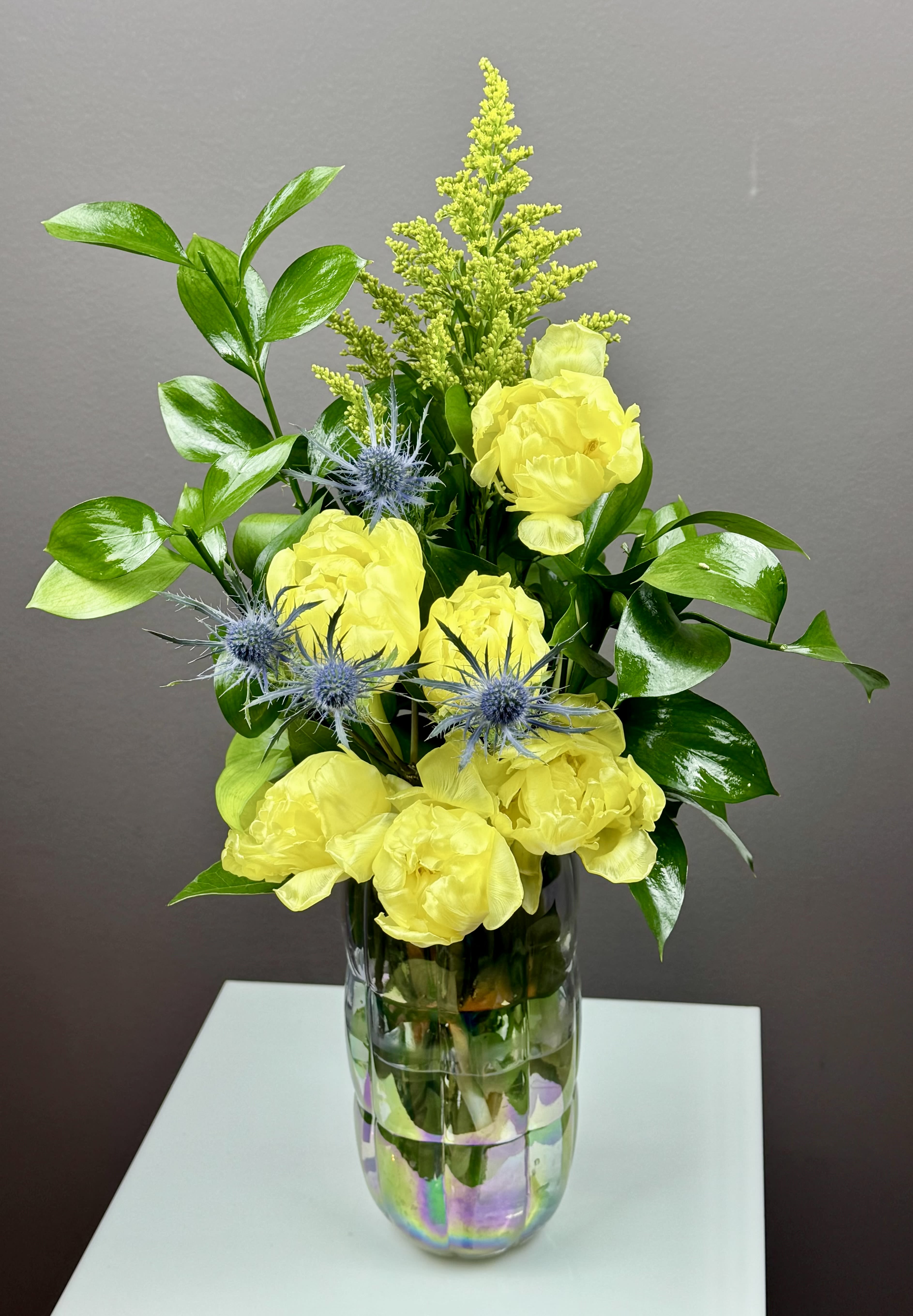 Golden Hour Glow - Illuminate any space with this vibrant, high-energy design that balances bold color with architectural interest. This arrangement features lush, sun-drenched blooms with ruffled, multi-layered petals that evoke the fullness of a summer garden.  The brilliant golden tones are artfully contrasted by spiky, indigo-blue accents, creating a sophisticated color palette that feels both edgy and elegant. A plume of textured, lime-green filler adds height and a touch of wildness, while deep, emerald-green foliage provides a glossy, polished frame.  Housed in a stunning iridescent ribbed glass vase that reflects a spectrum of colors, this piece is more than just a bouquet—it’s a modern statement. Perfect for a striking centerpiece or a thoughtful gift to celebrate a special moment.  Product Details:   Dimensions: Approximately 18 inches high by 9 inches wide.   Color Palette: Sunbeam Yellow, Electric Blue, and Deep Emerald.   Vibe: Modern, Textured, and Energetic.   Vase: Premium iridescent glass with a classic ribbed texture.   Ideal for: Birthdays, Thank-yous, or brightening a contemporary home.