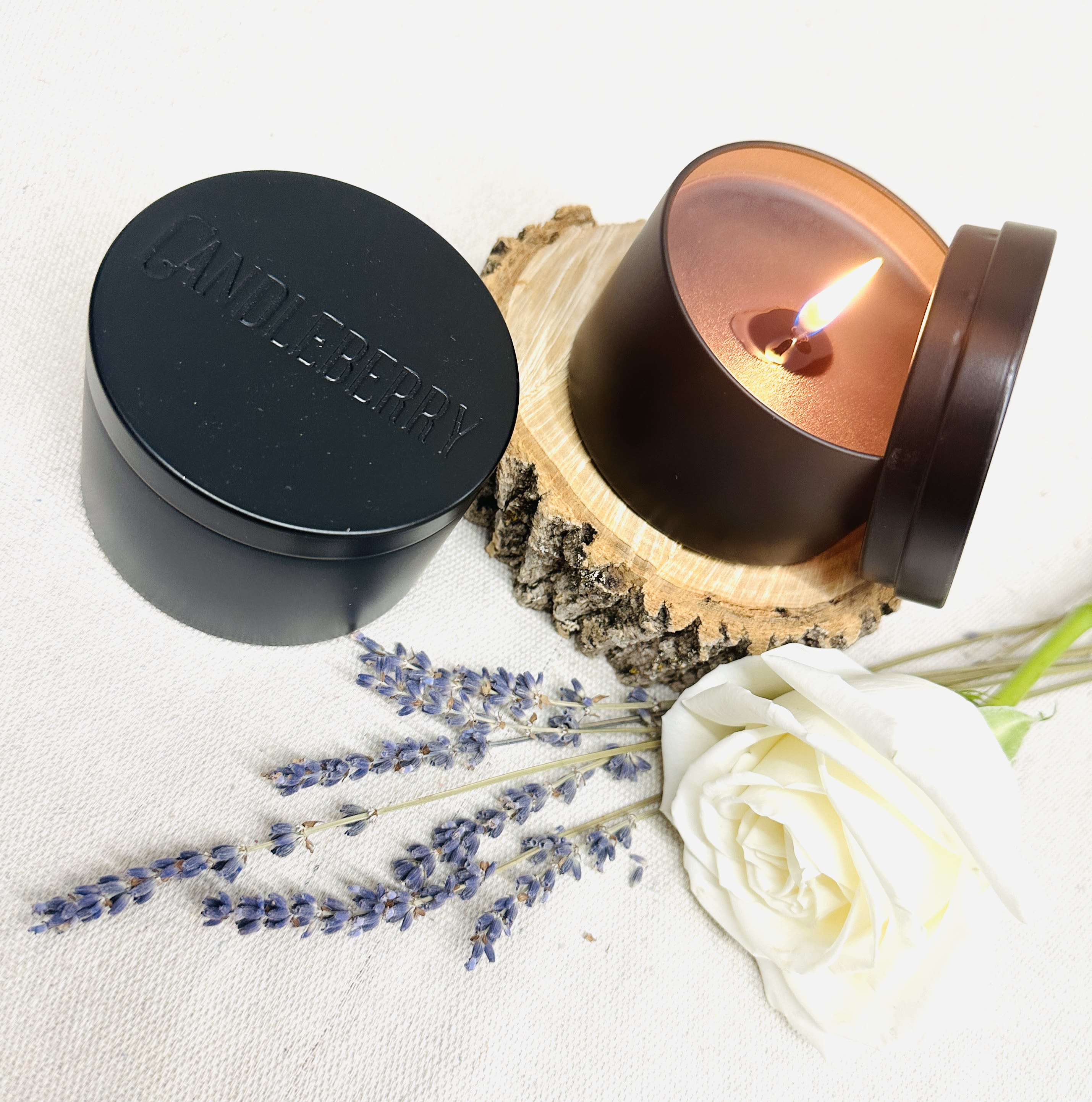 Lit candle in a black tin beside a white rose and lavender sprigs