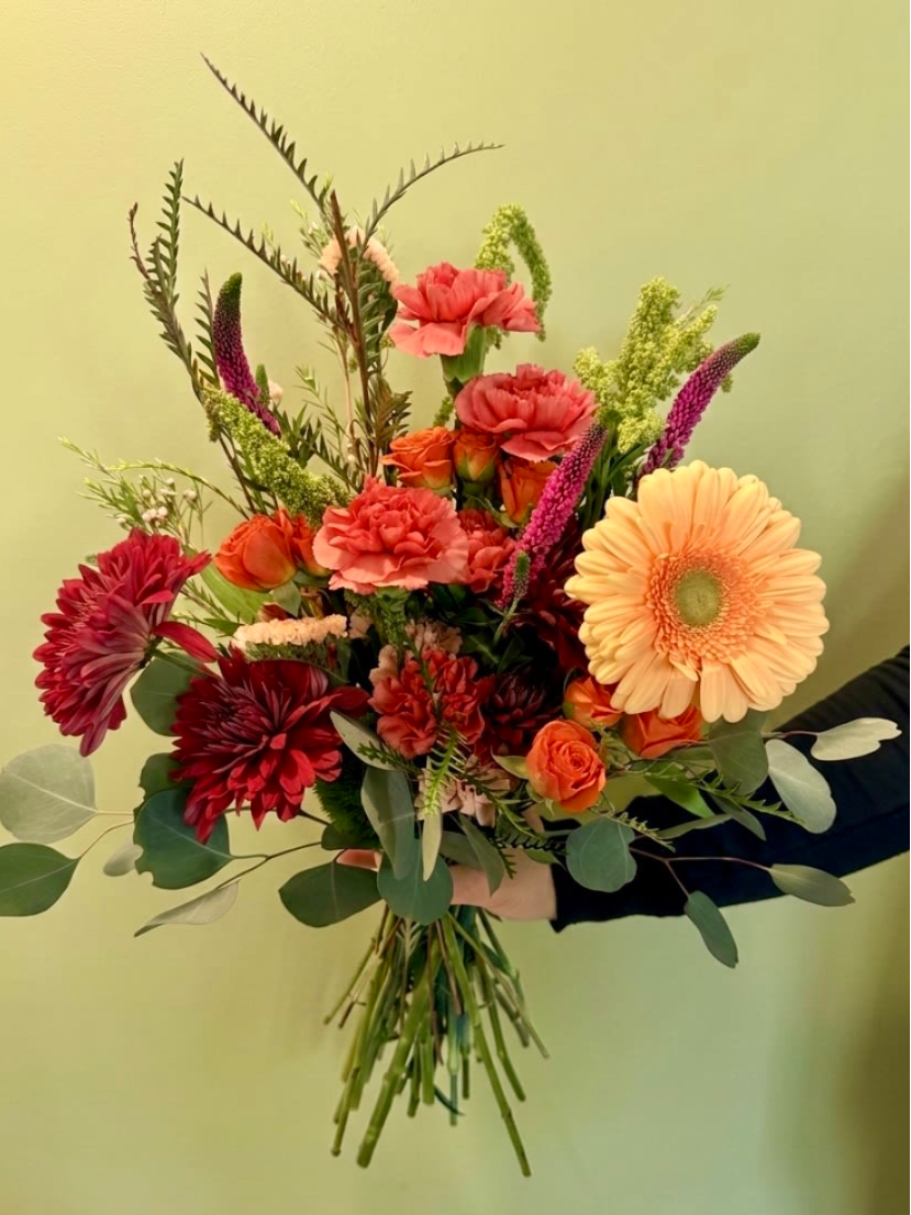Hand-tied bouquet of orange gerbera, red mums, carnations, and roses with tall filler stems, held against a pale wall