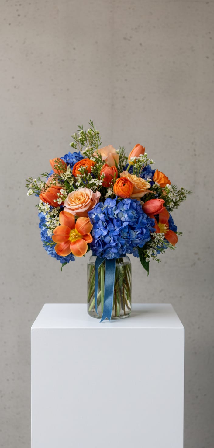 Bouquet of blue hydrangeas, orange tulips, and peach roses in a glass vase