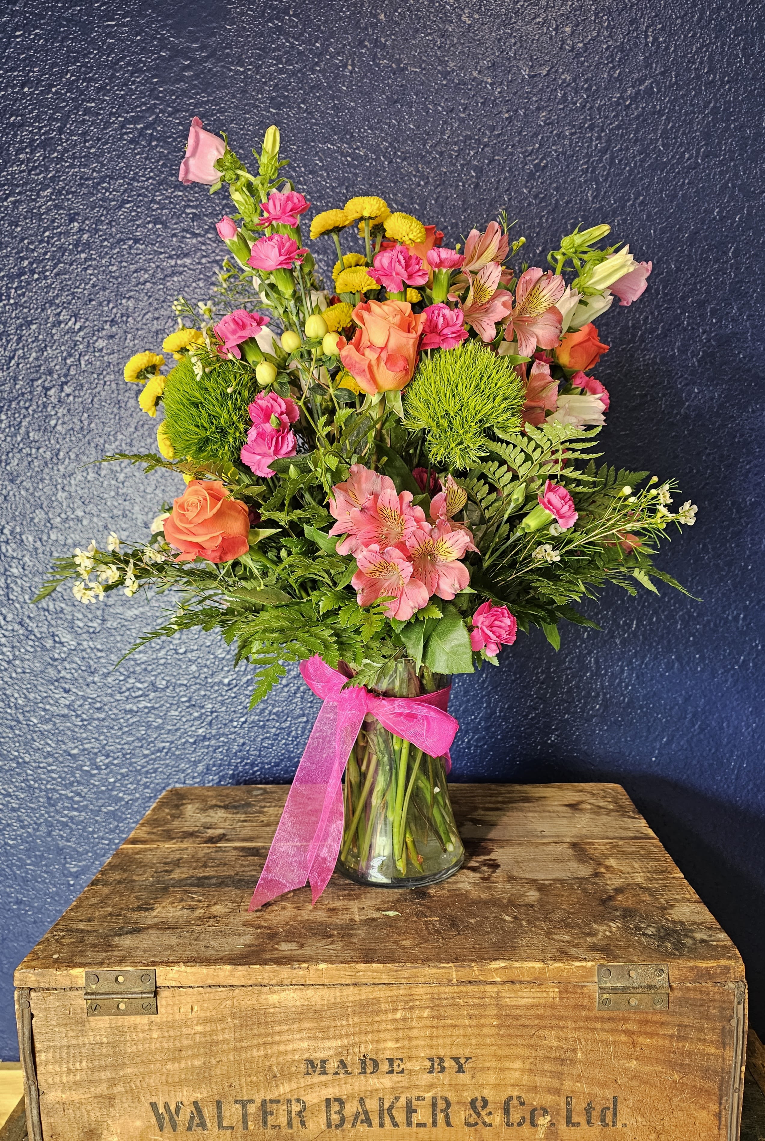 Mixed bouquet of pink, orange, and yellow flowers in a glass vase with a pink ribbon