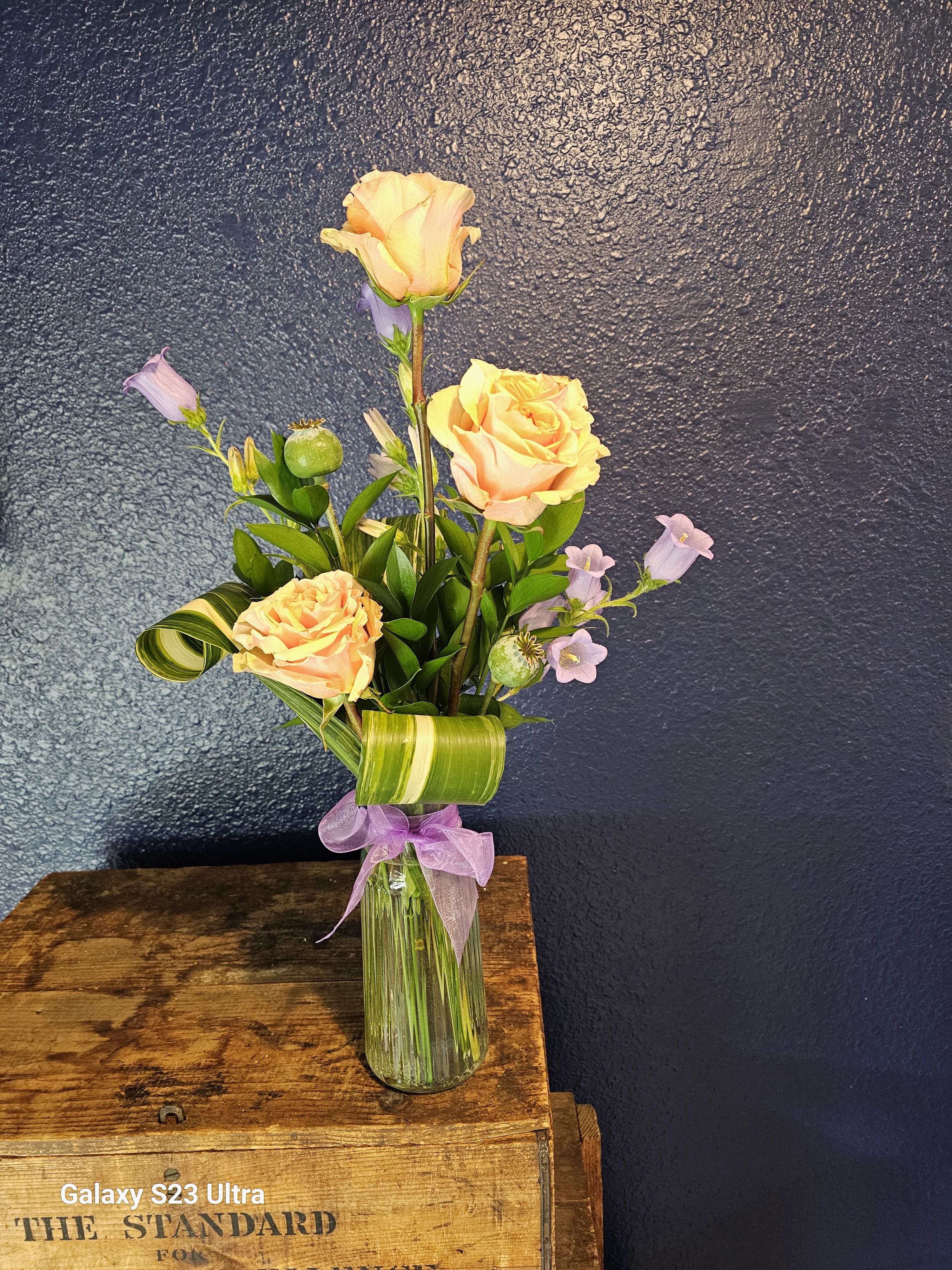 Peach roses and lavender bellflowers in a clear glass vase with a purple ribbon
