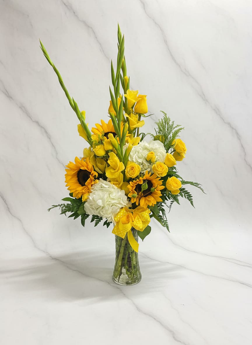 Blooming Sunshine Vase - A bright arrangement featuring yellow sunflowers, roses, spray roses, and gladiolas, accented with white hydrangea in a tall glass vase. Designed with a front-facing style.