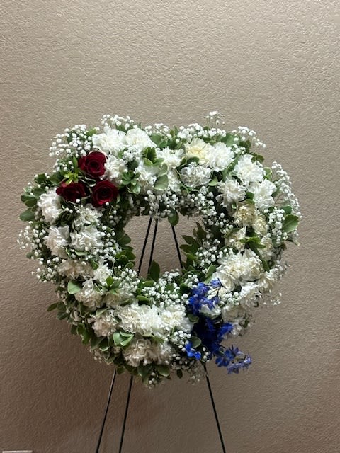 Heart-shaped floral wreath with white flowers, red roses, and blue accents on a stand