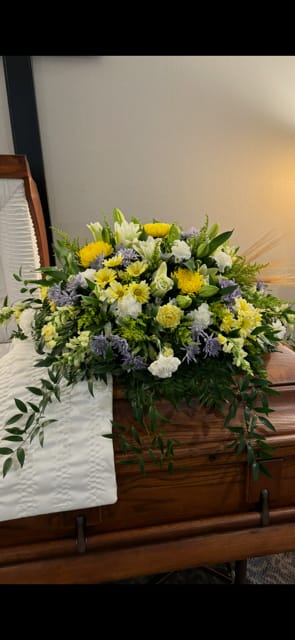 Large floral spray on a wooden casket with yellow, white, and lavender flowers.