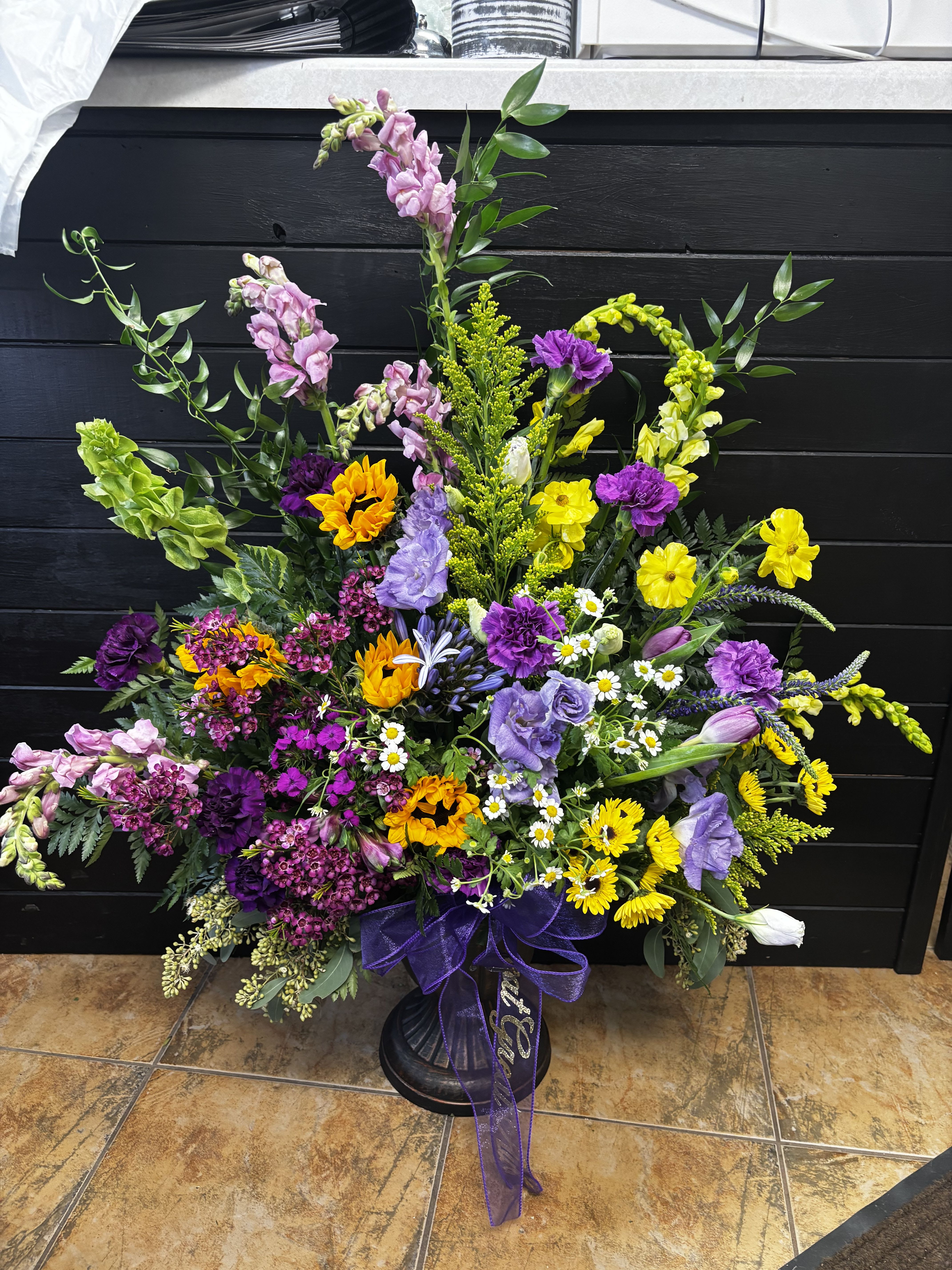 Sunlit Meadow Tribute - The Sunlit Meadow Tribute is a vibrant and uplifting sympathy arrangement inspired by the beauty of a blooming wildflower meadow. Cheerful sunflowers and golden blossoms shine among rich purple flowers, soft lavender tones, and delicate daisies. Tall snapdragons and flowing greenery add graceful height and movement, creating a natural garden-style display.