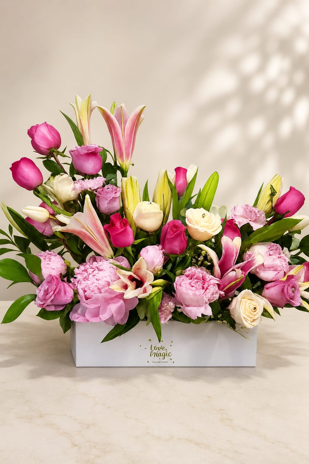 SUBLIME AMOR  - Soft pink lilies soaring above a bed of rounded roses and ruffled carnations give this flower arrangement an immediately romantic, upbeat presence - perfect as a birthday bouquet or a cheerful anniversary arrangement. The low, wide white rectangular box keeps the composition modern and gift-ready, so the person you're thinking of will feel seen the moment the lid lifts.  This arrangement mixes pink and cream roses with pale yellow and pink lilies and green accents for a fresh, layered look. The lilies rise above denser clusters of carnations and roses, creating height and movement while the white and yellow touches soften the stronger pinks. The compact box design makes it easy to place on a dining table or desk, presenting a polished, abundant display.  Send this as a heartfelt everyday gift, a congratulations bouquet, or a bright pick-me-up; it's the kind of arrangement that reads as both celebratory and sincere. The low rectangular box keeps stems neatly arranged - swap water daily and recut stems as needed to extend freshness.