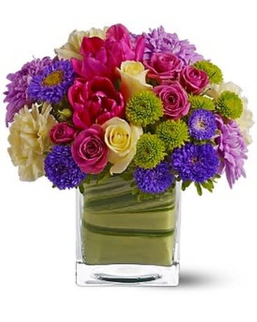 Mixed bouquet of pink, purple, and cream flowers in a clear square vase