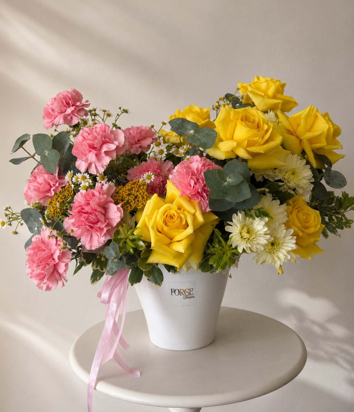 Pink carnations and yellow roses arranged in a white bucket vase with a pink ribbon.