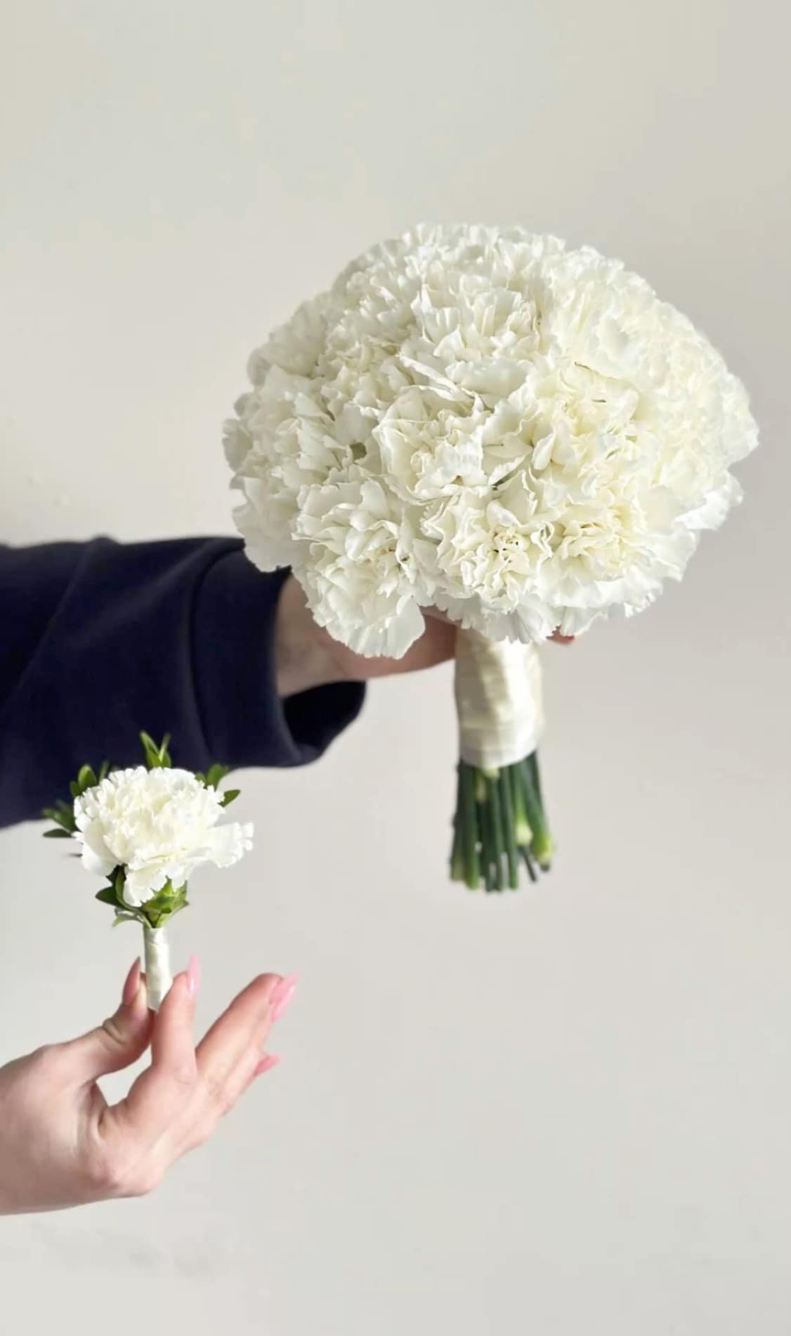 White carnation bouquet and matching boutonniere held against a plain background