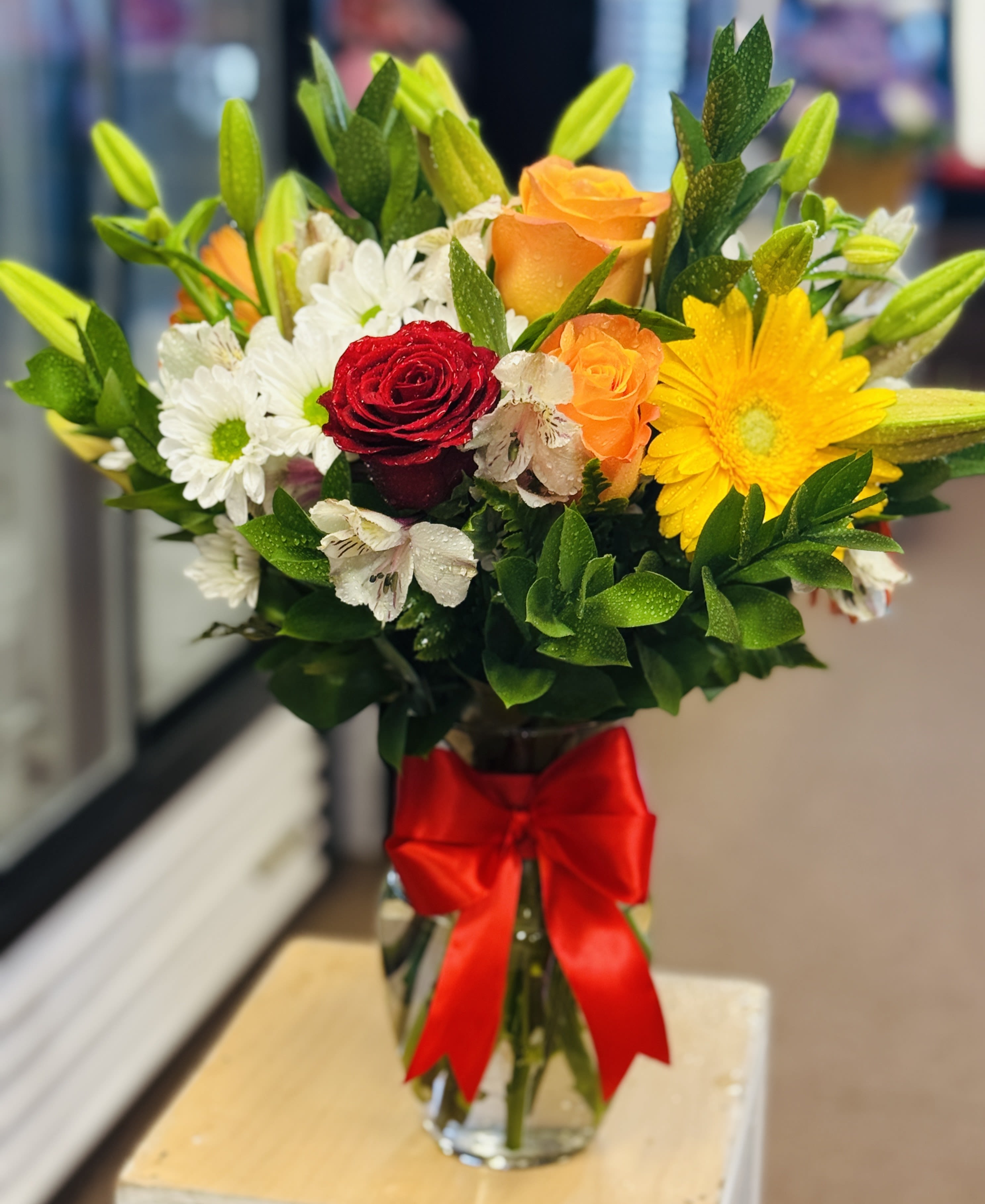 Bouquet of roses, daisies, and lilies in a glass vase with a red ribbon