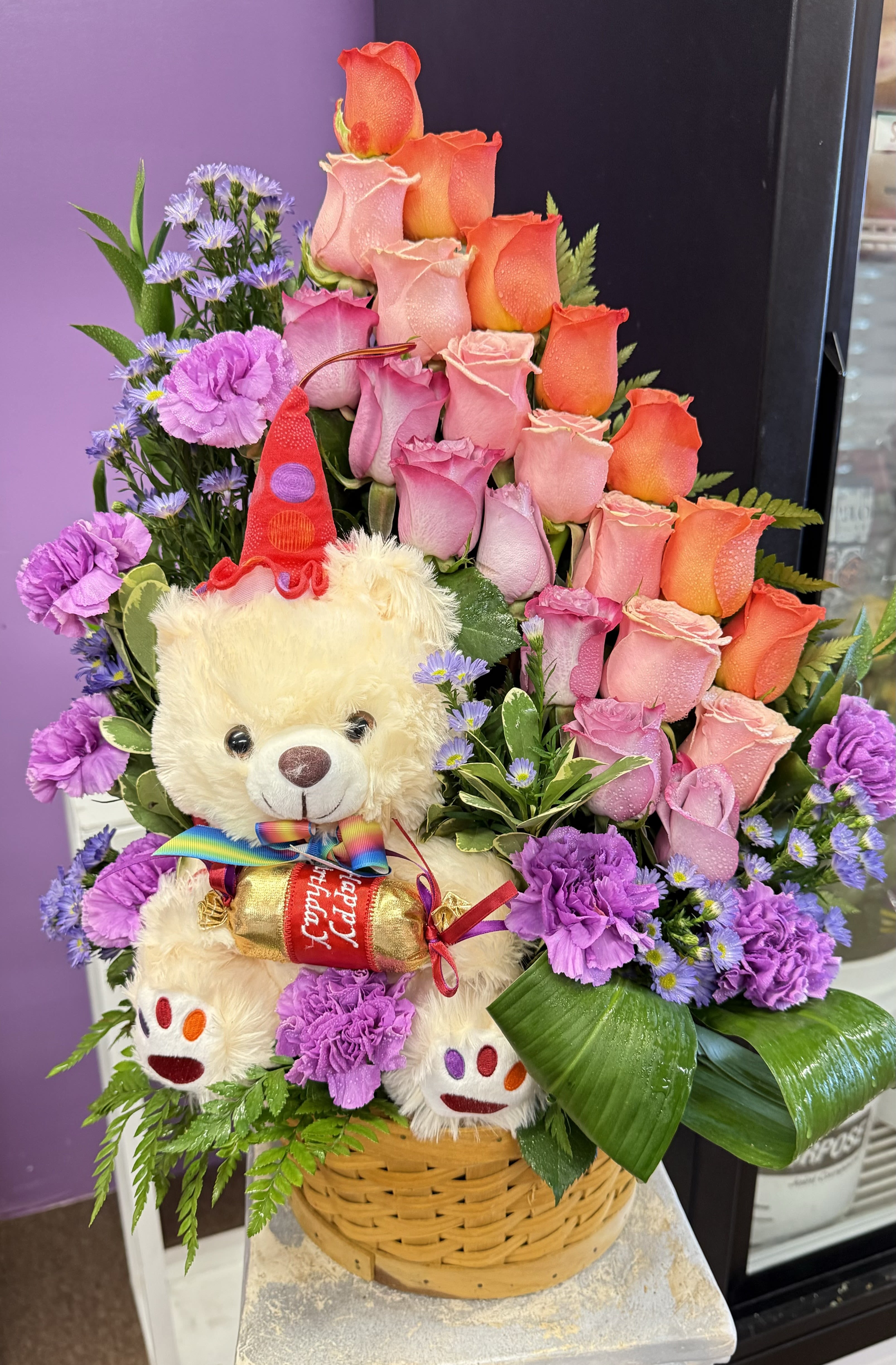 Basket arrangement of pink and orange roses with a teddy bear holding candy