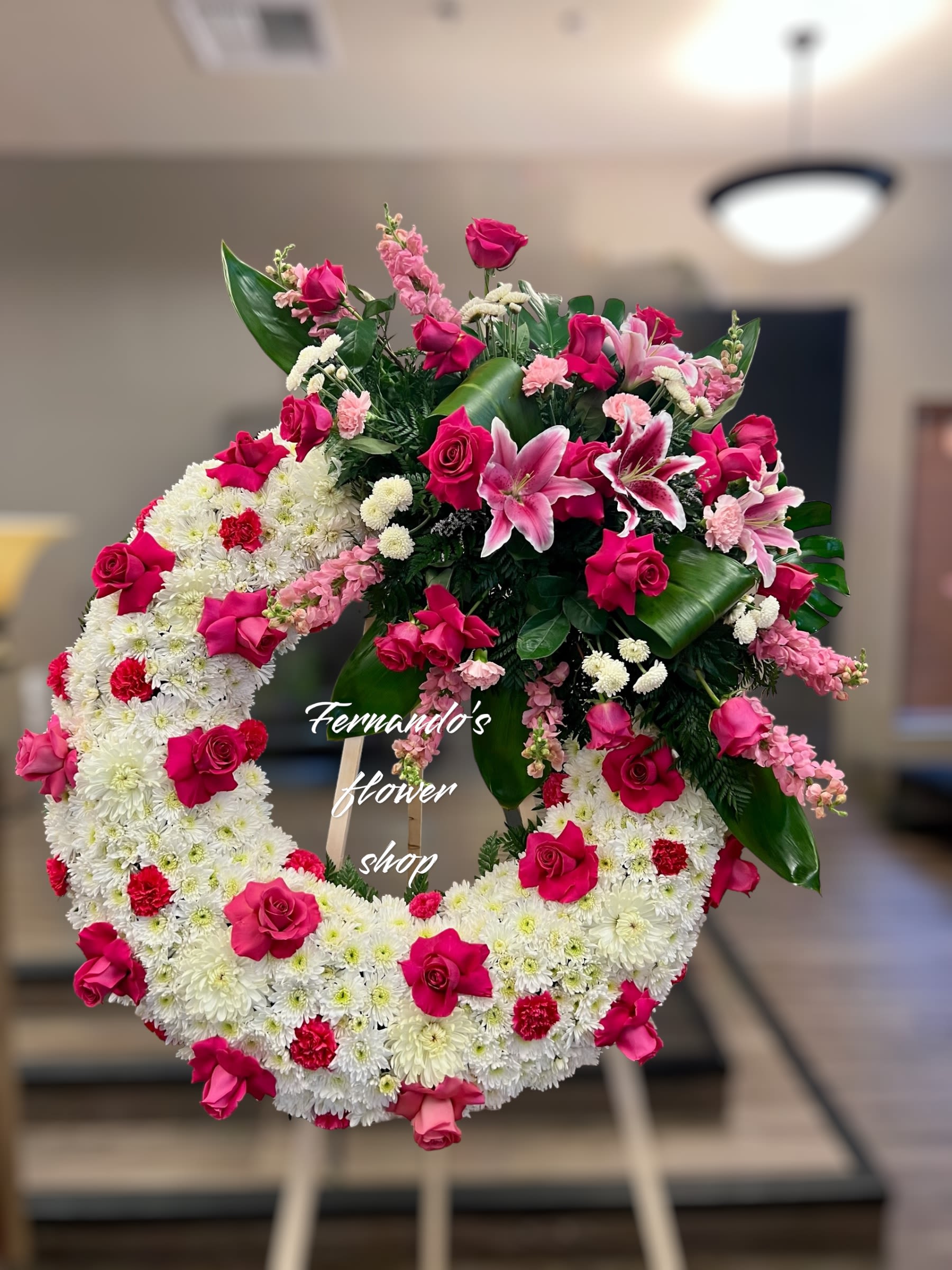 Blooming - “A magnificent funeral wreath featuring a blend of pure white chrysanthemums as the foundation, symbolizing peace and eternal rest. Bold red and pink roses, paired with delicate pink lilies, cascade gracefully from the top, creating a striking focal point. The vibrant flowers are complemented by lush greenery and soft pink accents, adding elegance and warmth. This arrangement is a timeless expression of love, honor, and heartfelt condolences, offering a beautiful tribute to a cherished loved one. ️”