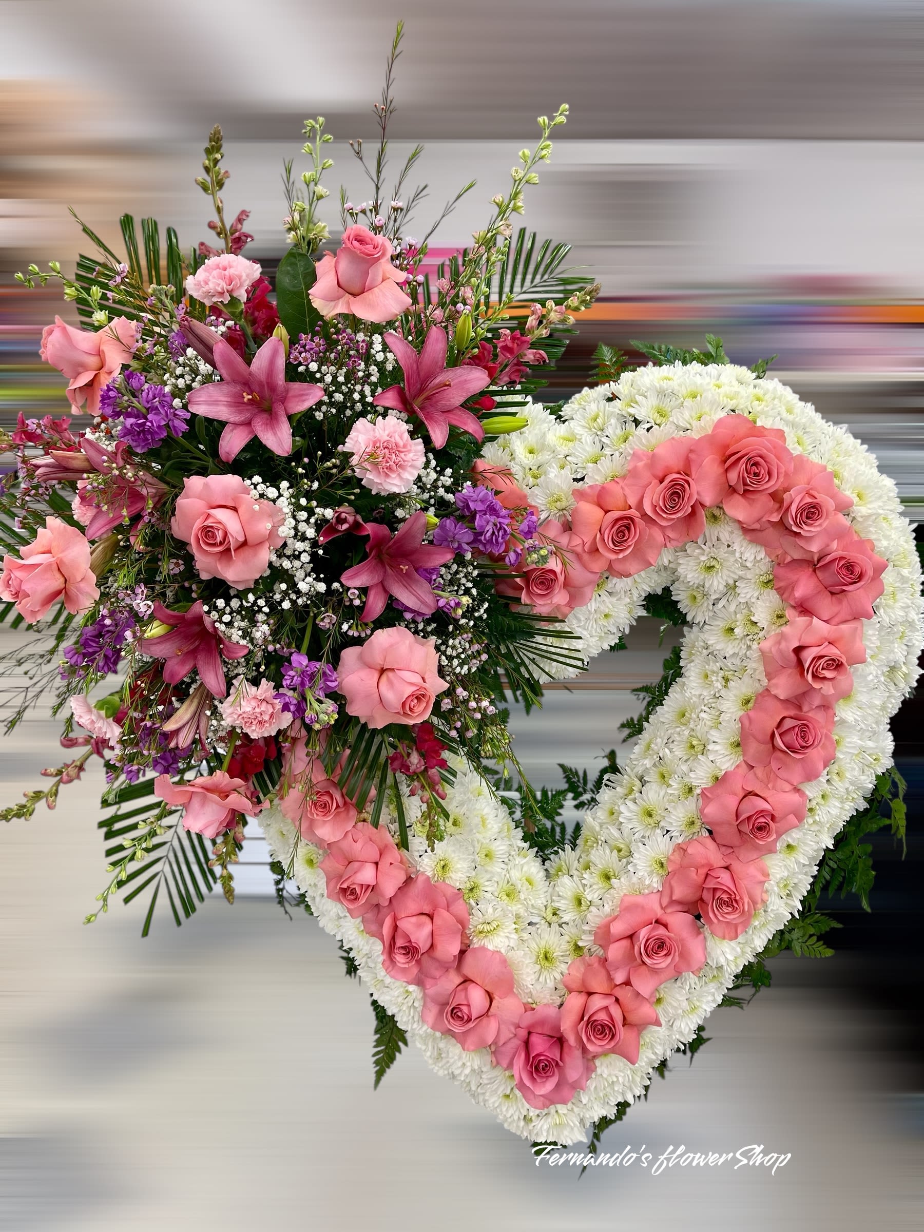 Loving Tribute  - Express love and heartfelt remembrance with this stunning heart-shaped floral arrangement. Crafted with a base of pure white chrysanthemums, symbolizing peace and purity, the heart is beautifully outlined with soft pink roses, representing love, admiration, and gratitude. A lush spray of pink lilies, roses, carnations, and purple blooms adorns the side, adding vibrant color and elegance. This thoughtful tribute is perfect for honoring a cherished soul and celebrating a life filled with love.