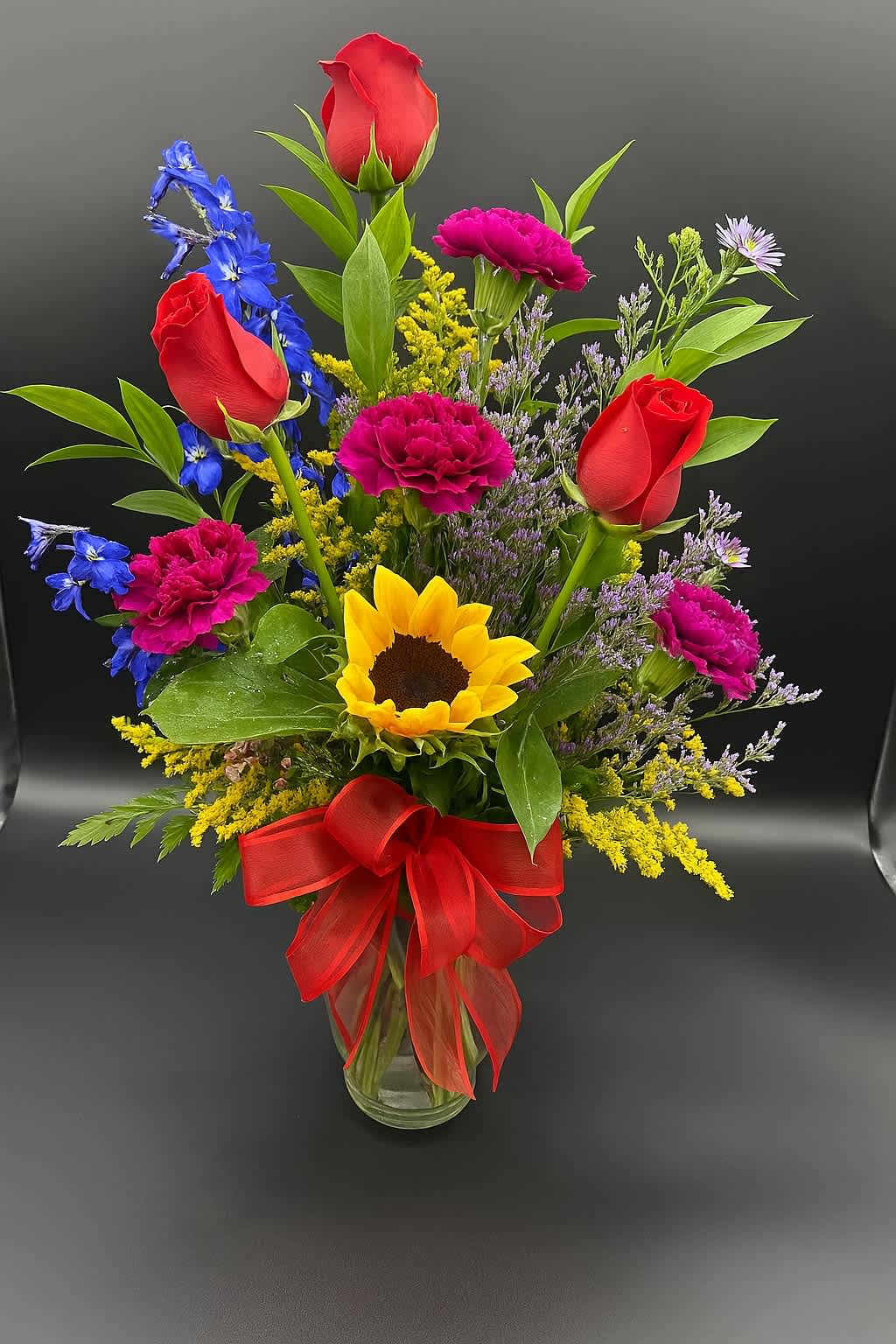 Special Occasion - Bright, bold, and full of cheer! This vibrant vase features a joyful mix of red roses, sunflowers, hot pink carnations, and blue delphiniums—perfect for lifting spirits or celebrating any special occasion.