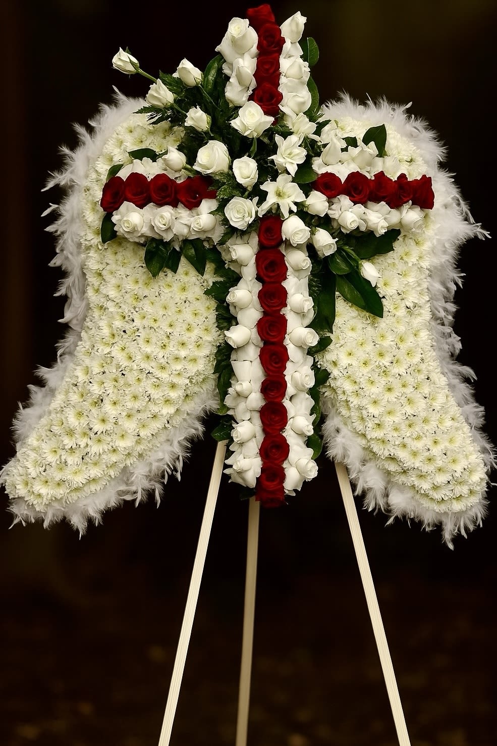 Wings of Peace 1 - This magnificent floral arrangement is a heartfelt tribute featuring angel wings and a cross design, symbolizing faith, love, and eternal peace. The wings are crafted with white chrysanthemums, representing purity and grace, bordered by delicate feather-like accents for a soft and heavenly touch. At the center, a striking cross is formed with alternating vibrant red and white roses, adding elegance and depth to the composition. Accented with lush greenery, this arrangement from “Fernando’s Flower Shop” is a beautiful expression of sympathy and devotion, perfect for honoring a loved one in a meaningful way. OVERSIZE PC