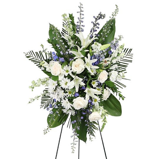 Breath Of The Heavens - A serene blue, white &amp; lavender sympathy standing spray designed with elegant white lilies and white roses, accented with soft lavender/blue delphinium and fresh white daisy mums, all framed with tropical greens and palm fronds. A peaceful tribute that honors a loved one with grace, comfort, and lasting love.