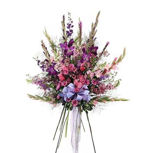 Elegance and Simplicity Standing Spray - An elegant standing floral arrangement featuring a vibrant blend of purple, lavender, and soft pink blooms. Accented with delicate greenery and tall, graceful stems, the design creates a sense of height and movement. A soft lavender ribbon bow at the center adds a gentle, heartfelt touch, making it a beautiful tribute suitable for expressing sympathy, remembrance.