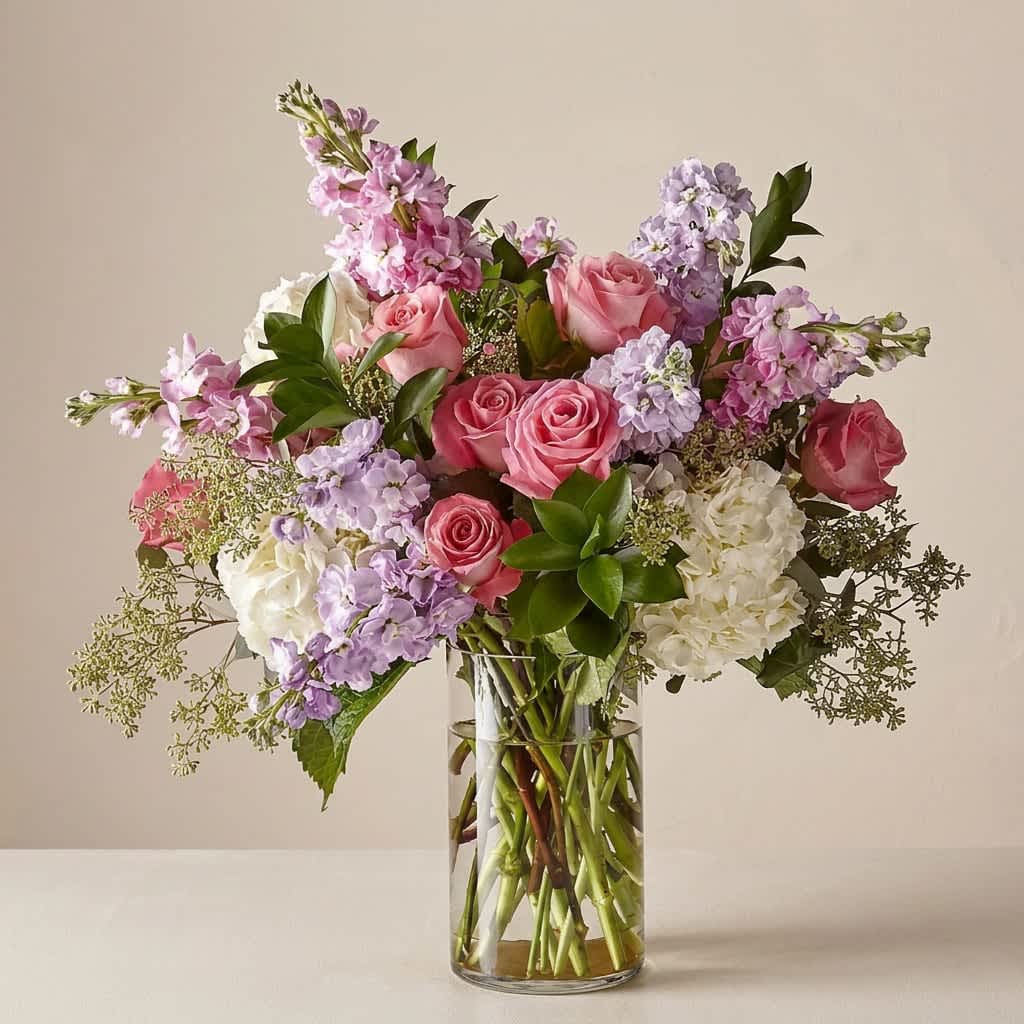 In the Garden (DL) - Experience the perfect blend of modern elegance and natural beauty with our stunning tall, In the Garden floral arrangement. This meticulously designed arrangement features a captivating combination of pink roses, stock, white hydrangeas, eucalyptus, and assorted greens, all expertly arranged in a sleek 6x10" leaf-lined glass cylinder vase.