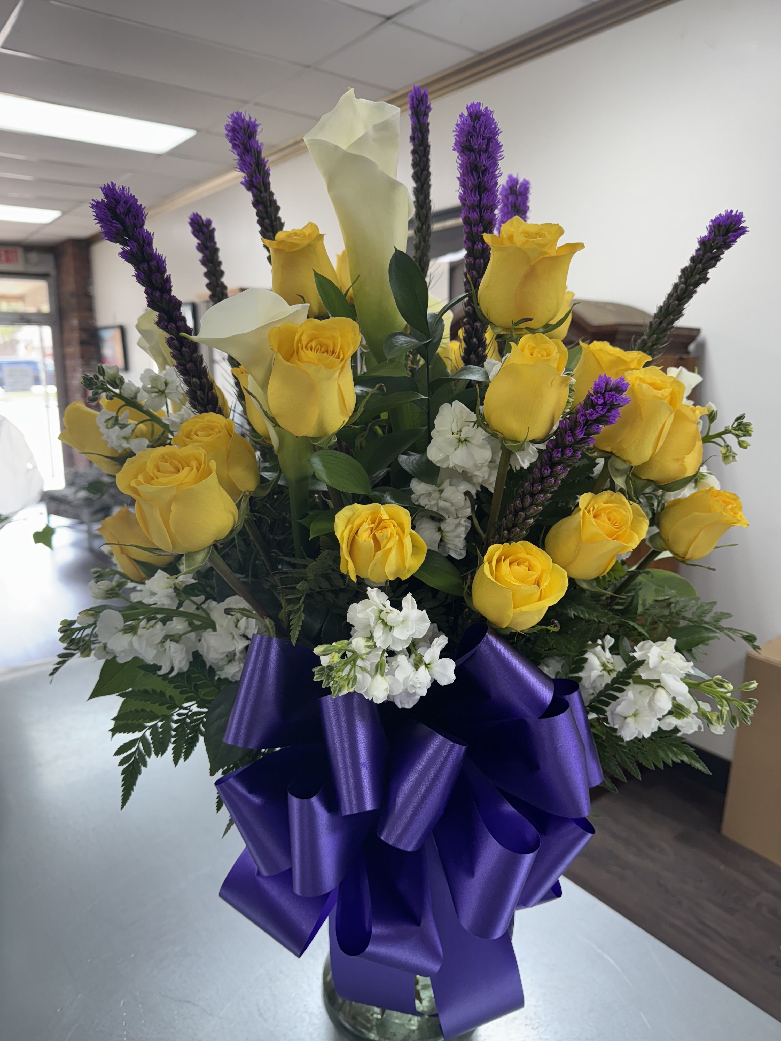 Yellow roses and white calla lilies in a glass vase with purple ribbon