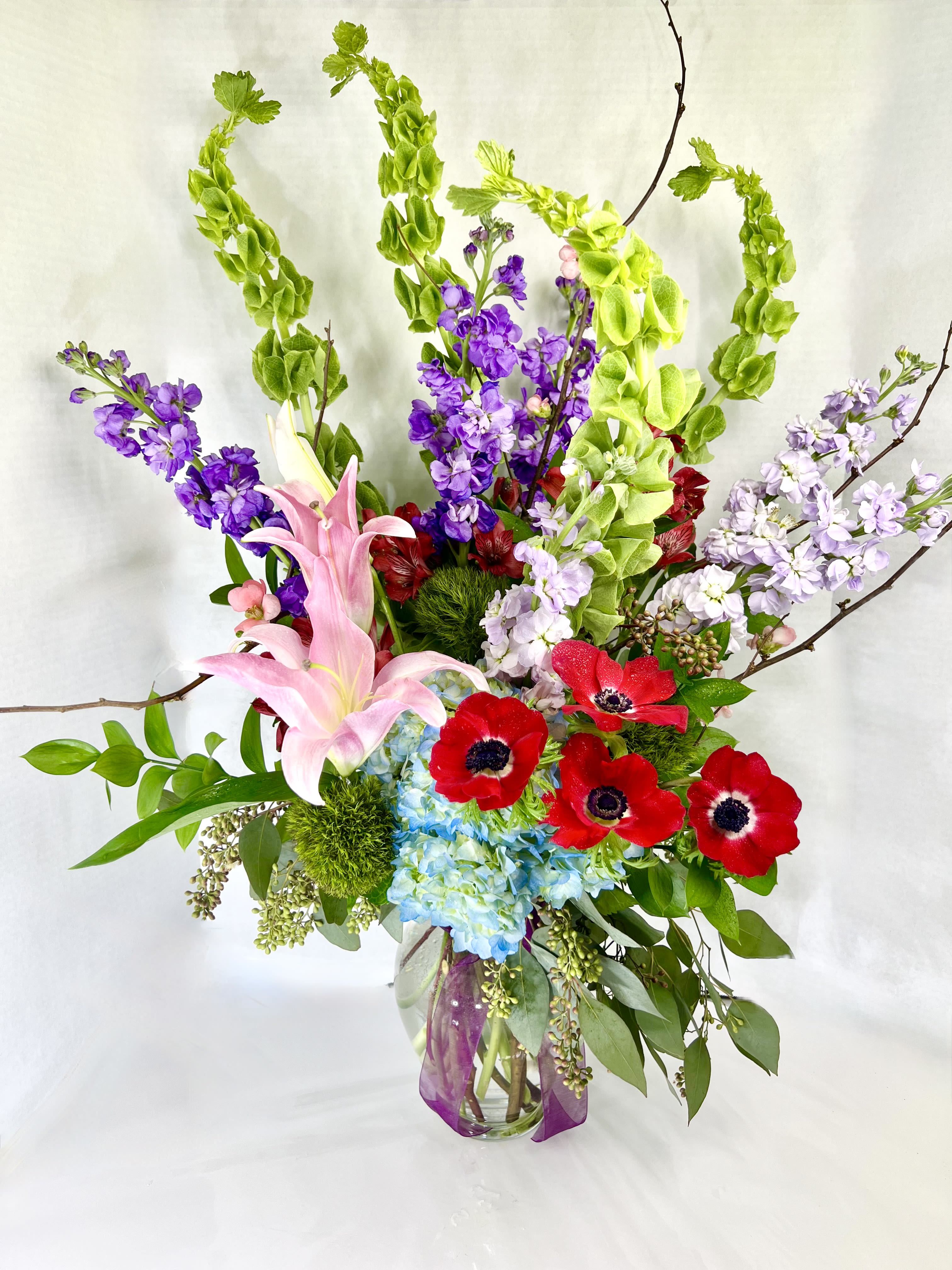 Tall bouquet of pink lilies, red anemones, and purple blooms in a glass vase